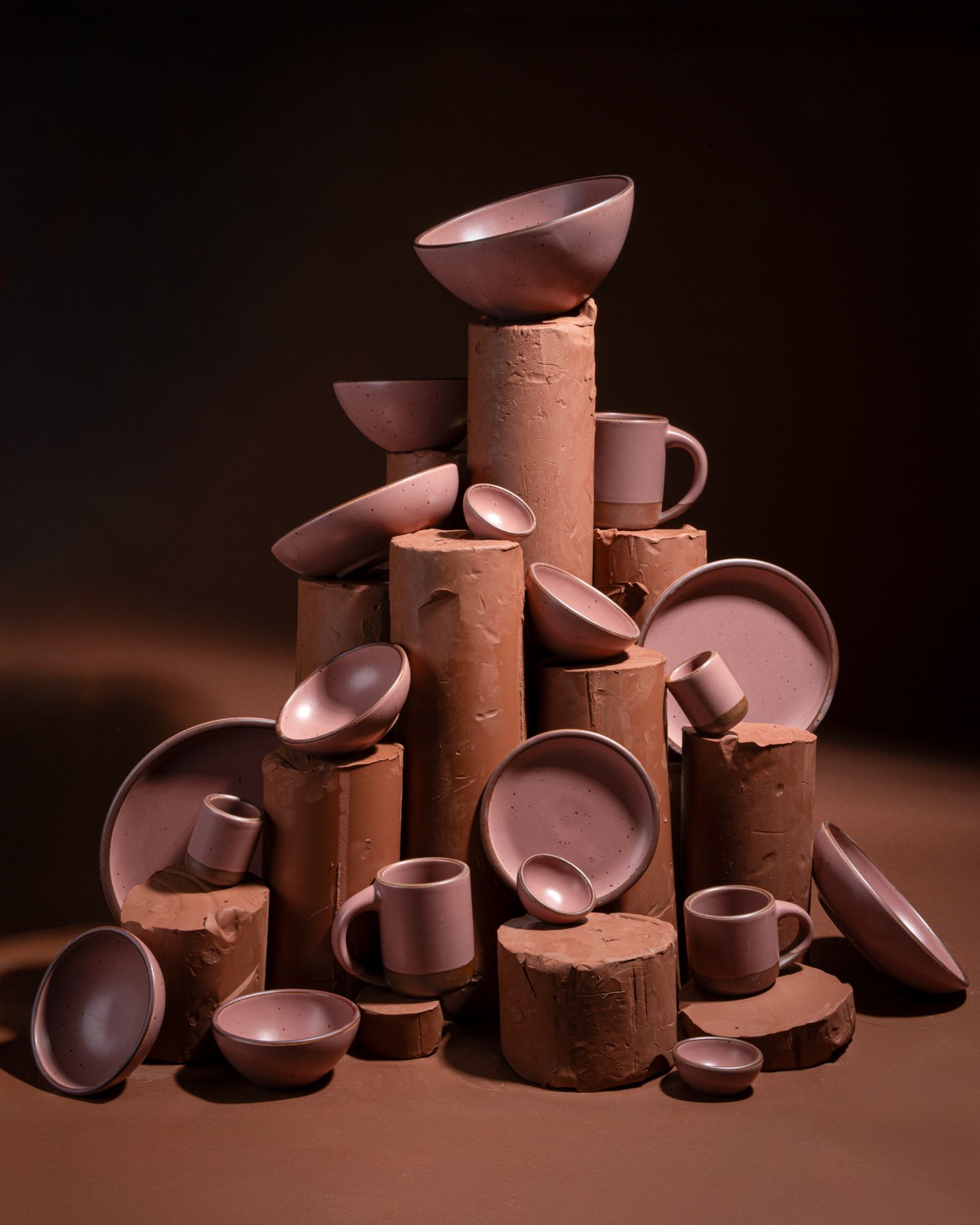 Dusty mauve pink ceramic bowls, plates, and mugs arranged on stacked clay cylinders against a dark studio background.