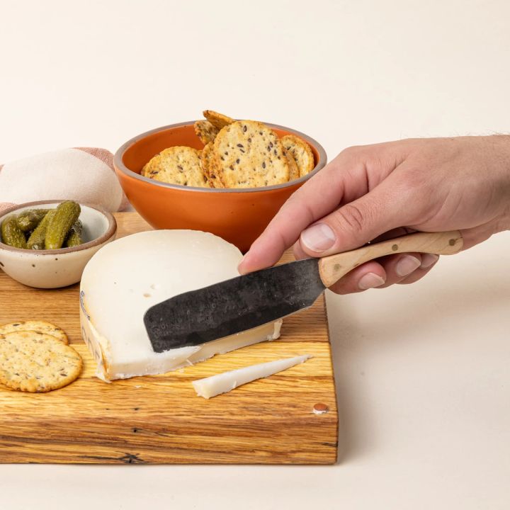 A hand holds a hand-forged cheese knife with a stainless steel rectangular blade and a maple wood handle. The blade is cutting some gouda, and surrounded by fixings for a charcuterie board.