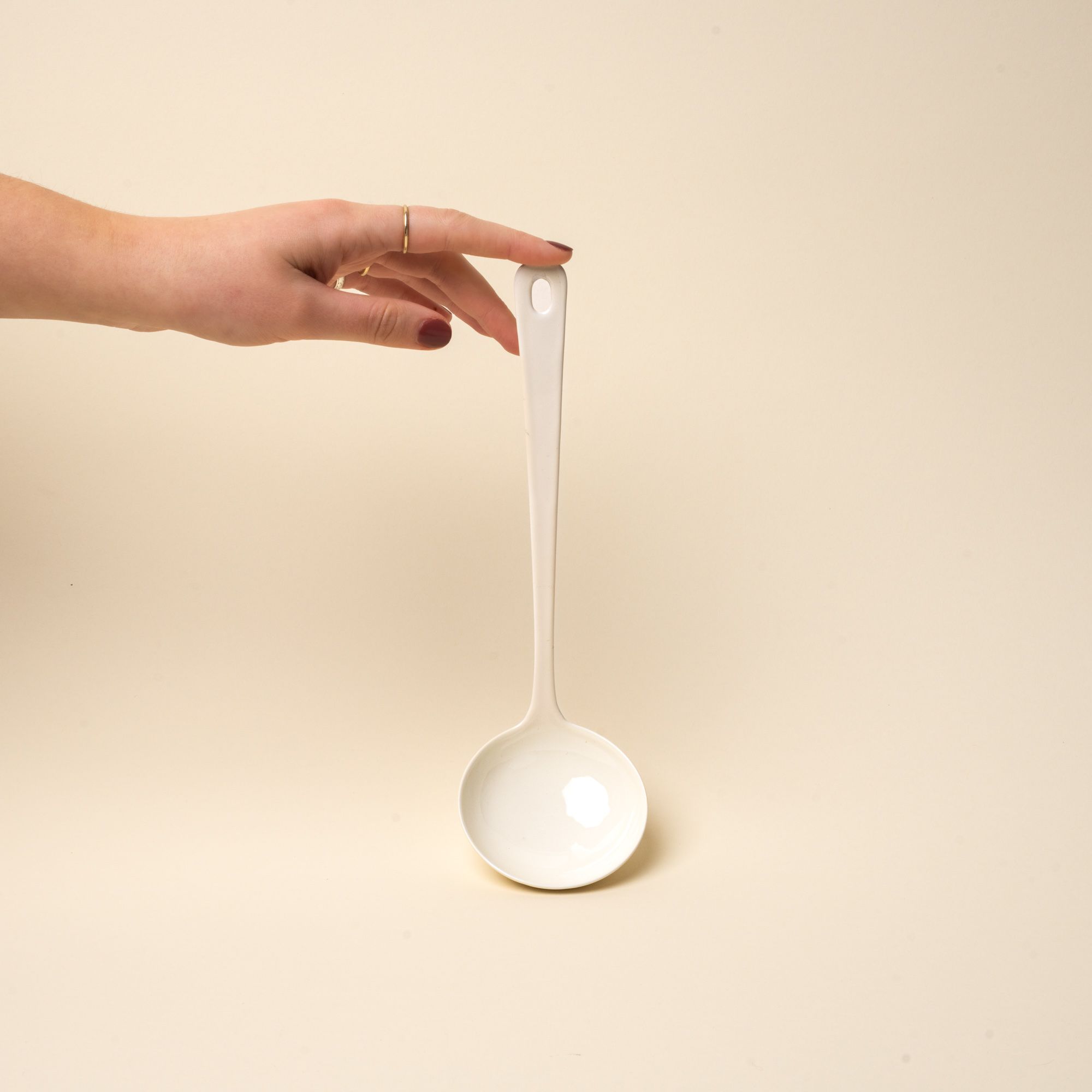 A hand props up a hand holds a white enamel ladle with a hole at the end of the handle for hanging.