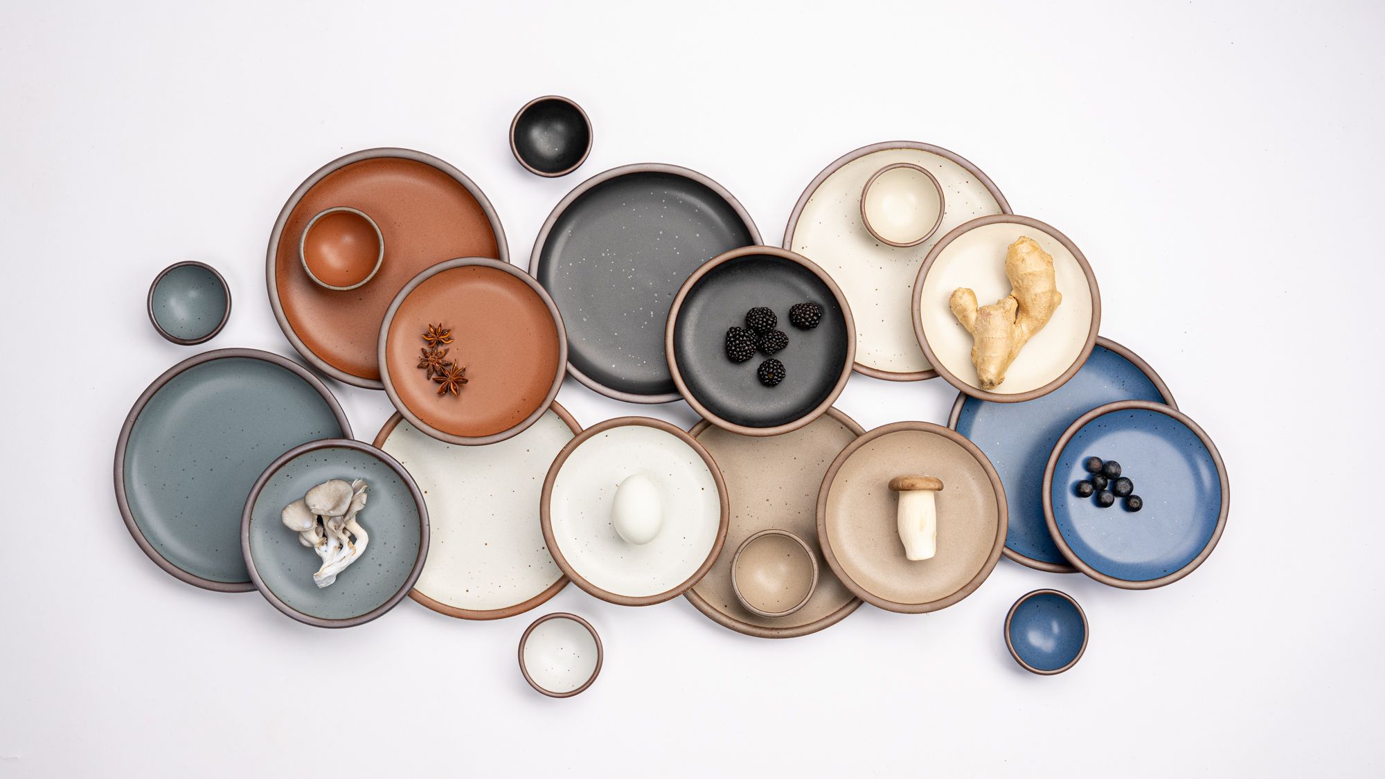 Various sizes of ceramic plates and tiny bowls in a rainbow of earthy colors are artfully laid out together with match ingredients against a white background.