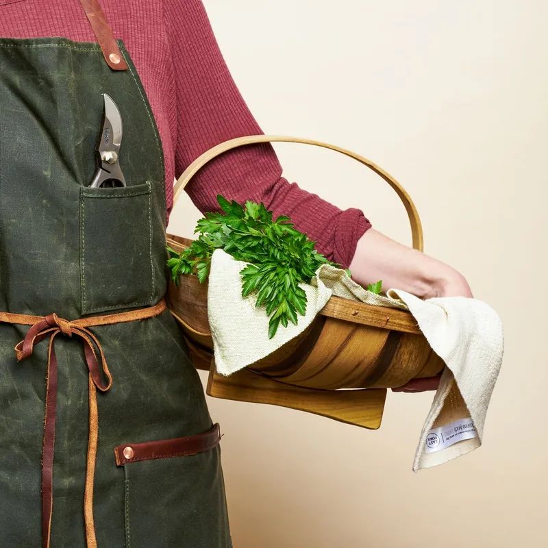 A hand holds a large myrtlewood trug to their hip and it is filled with a kitchen towel and a harvest