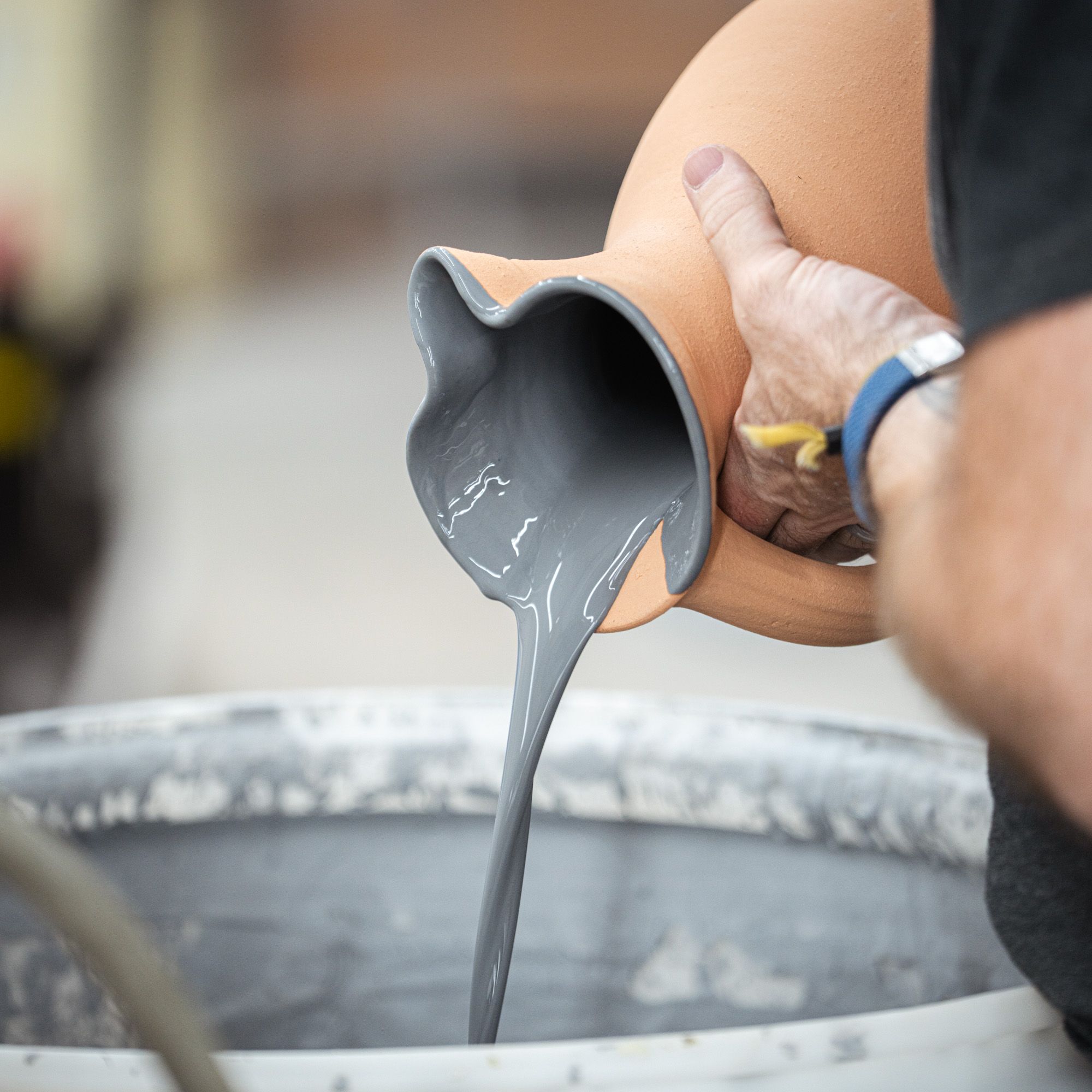Hands pouring gray glaze out of an unglazed pitcher into a large container.