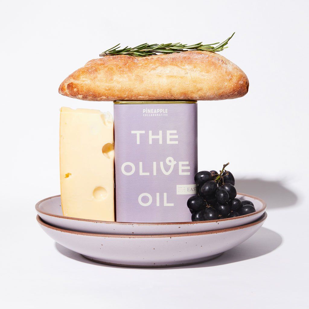 Two East Fork Coupes in Taro with a block of cheese, a can of olive oil with a purple label and a bunch of purple grapes with a loaf of bread with a spring of rosemary on top of the oil