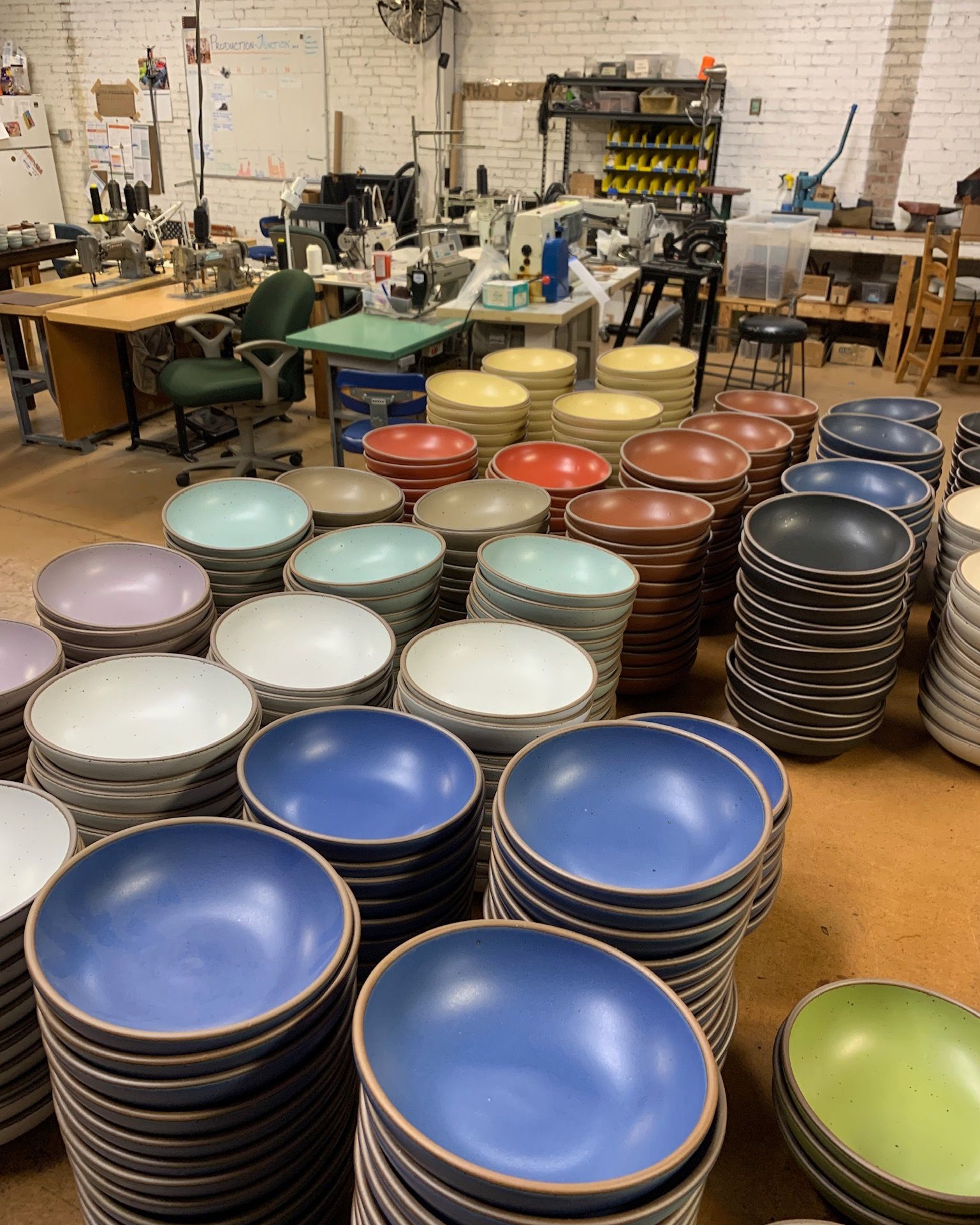 In a workshop setting, there are large stacks of ceramic bowls and plates in all different colors of the rainbow arranged on a table