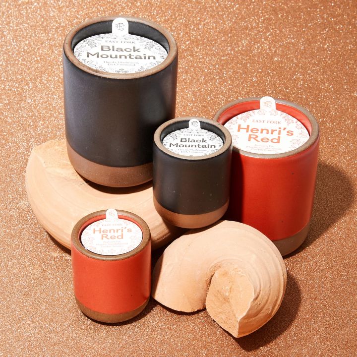 4 ceramic vessel with candles inside styled together on a warm brown background with 2 wood pieces. Two candles are in a orange-red color and the other two are in a dark charcoal color. On top of each is a packaging label sitting that reads "Black Mountain" or "Henri's Red"
