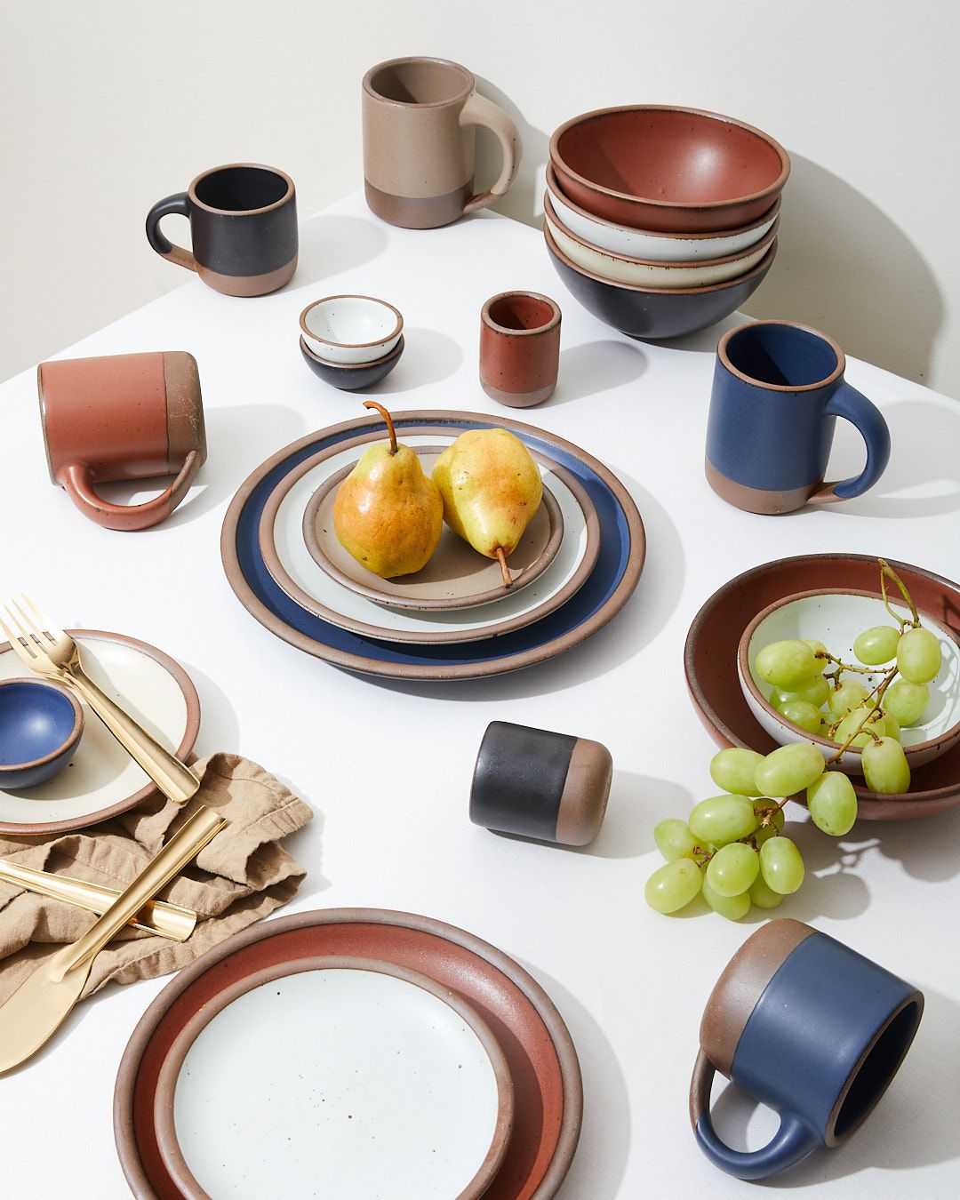 An artful arrangement on a table of ceramic cups, mugs, bowls, and plates in white, terracotta, blue, tan, and black colors with fruit and silverware.