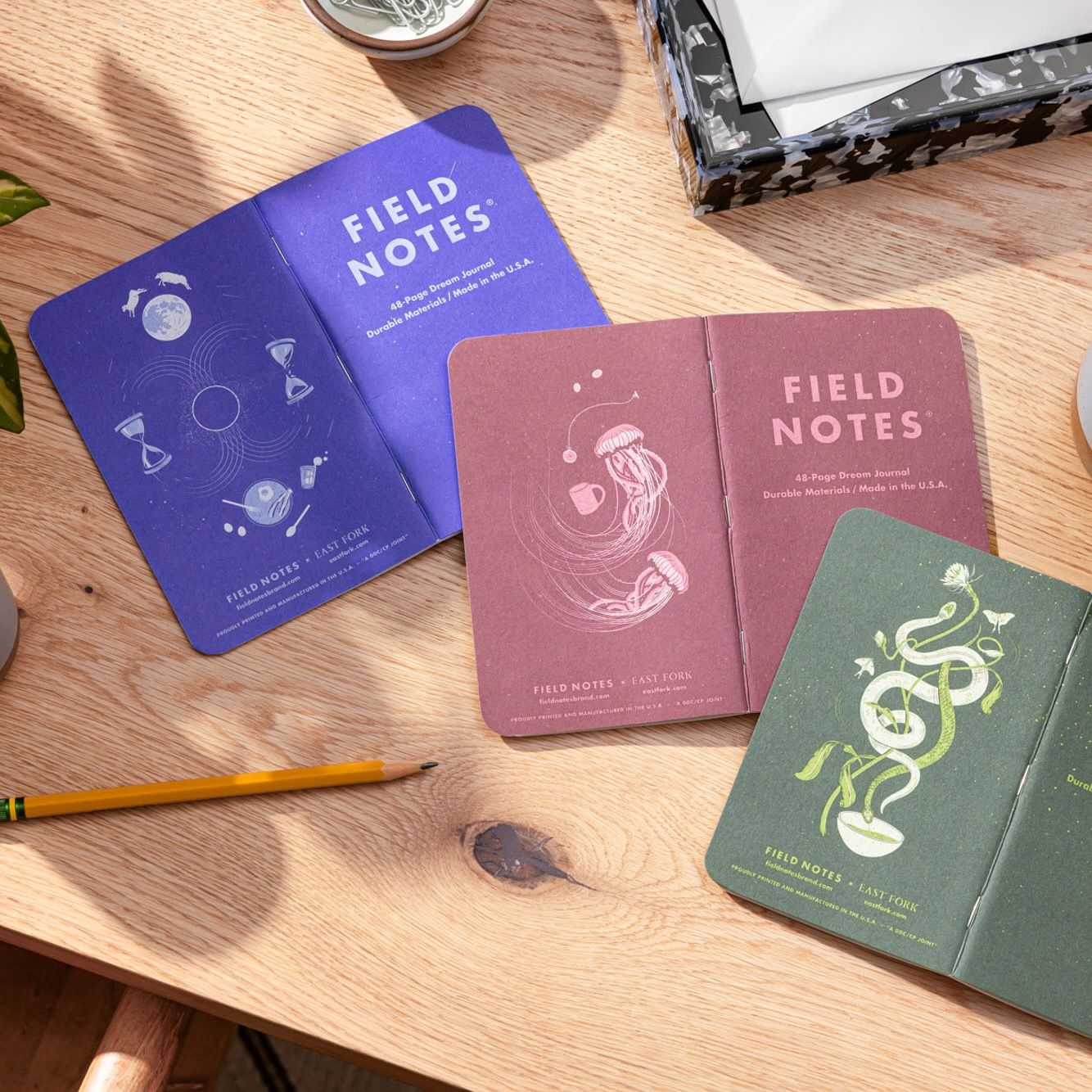 A 3 pack of Field Notes Journal are folded facing down on a desk featuring 3 illustrated back covers in a lapis blue, a plum, and an earthy green.