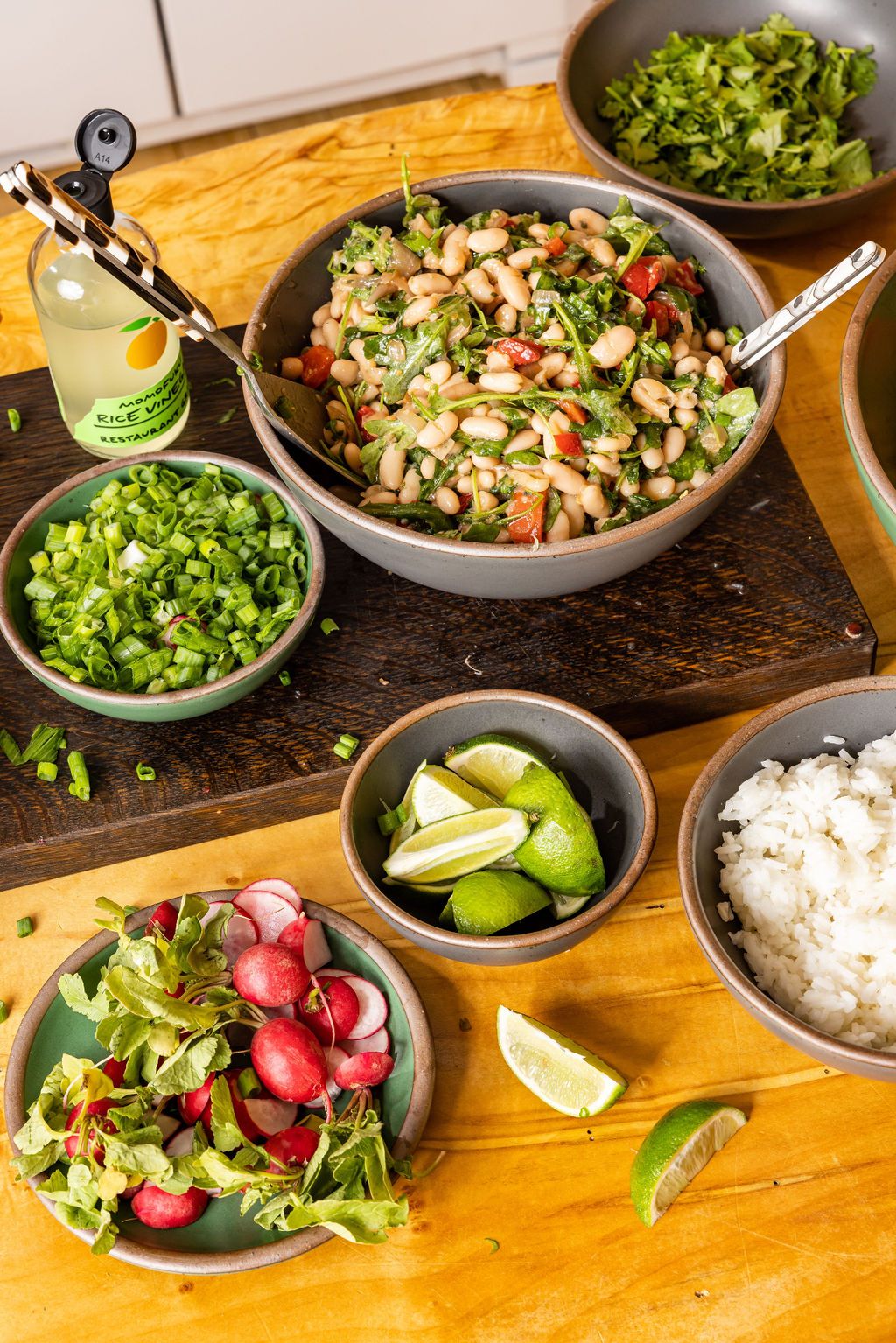 A large bowl with a bean salad, a smaller bowl next to it with green onions, another bowl with limes and a bowl with radishes