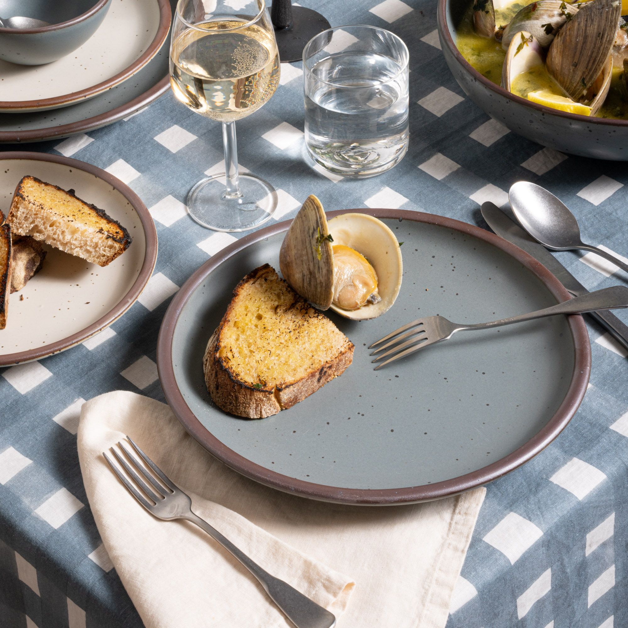 Clams and toast on a blue-grey dinner plate with silverware on a blue-grey crosshatch tablecloth