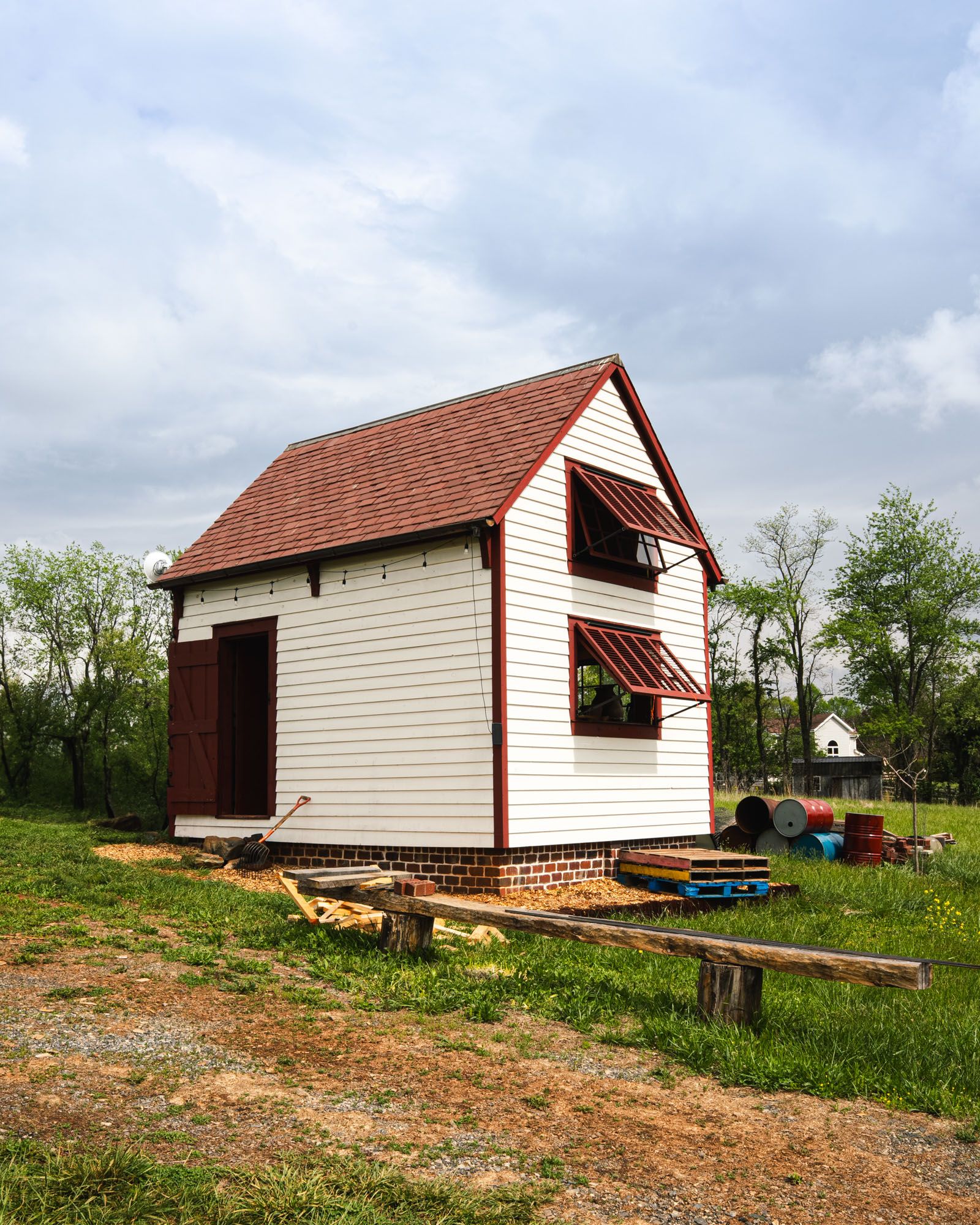 Small white building with red trim and open awning windows, set on grass with tools, barrels, and pallets nearby.