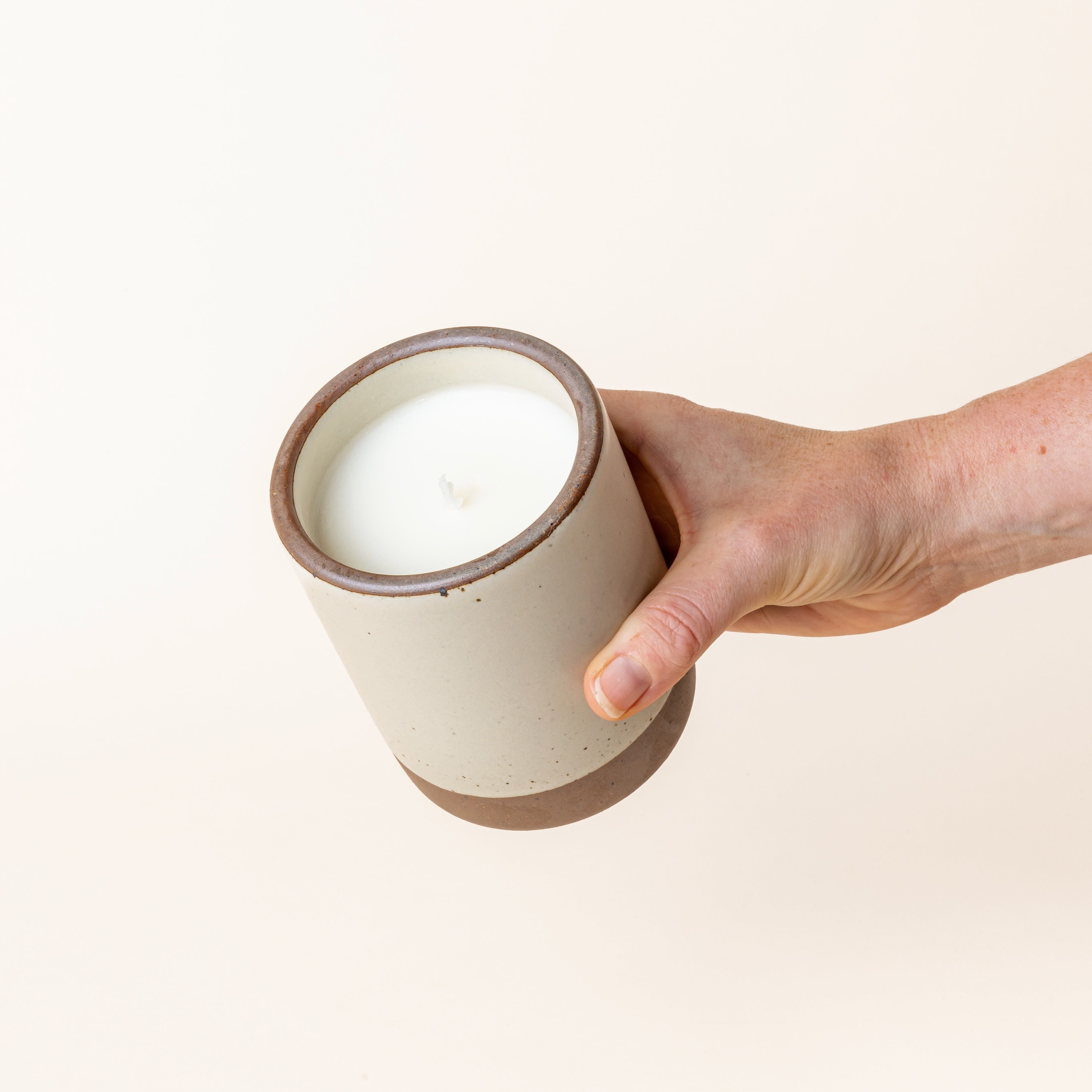 Hand holding a large ceramic vessel in a warm, tan-toned, off-white color with candle inside.