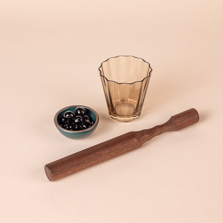 A long walnut wood cocktail muddler that tapers slightly towards the handle. Next to it is a short amber ridged lowball glass and a tiny teal ceramic bowl filled with cherries.