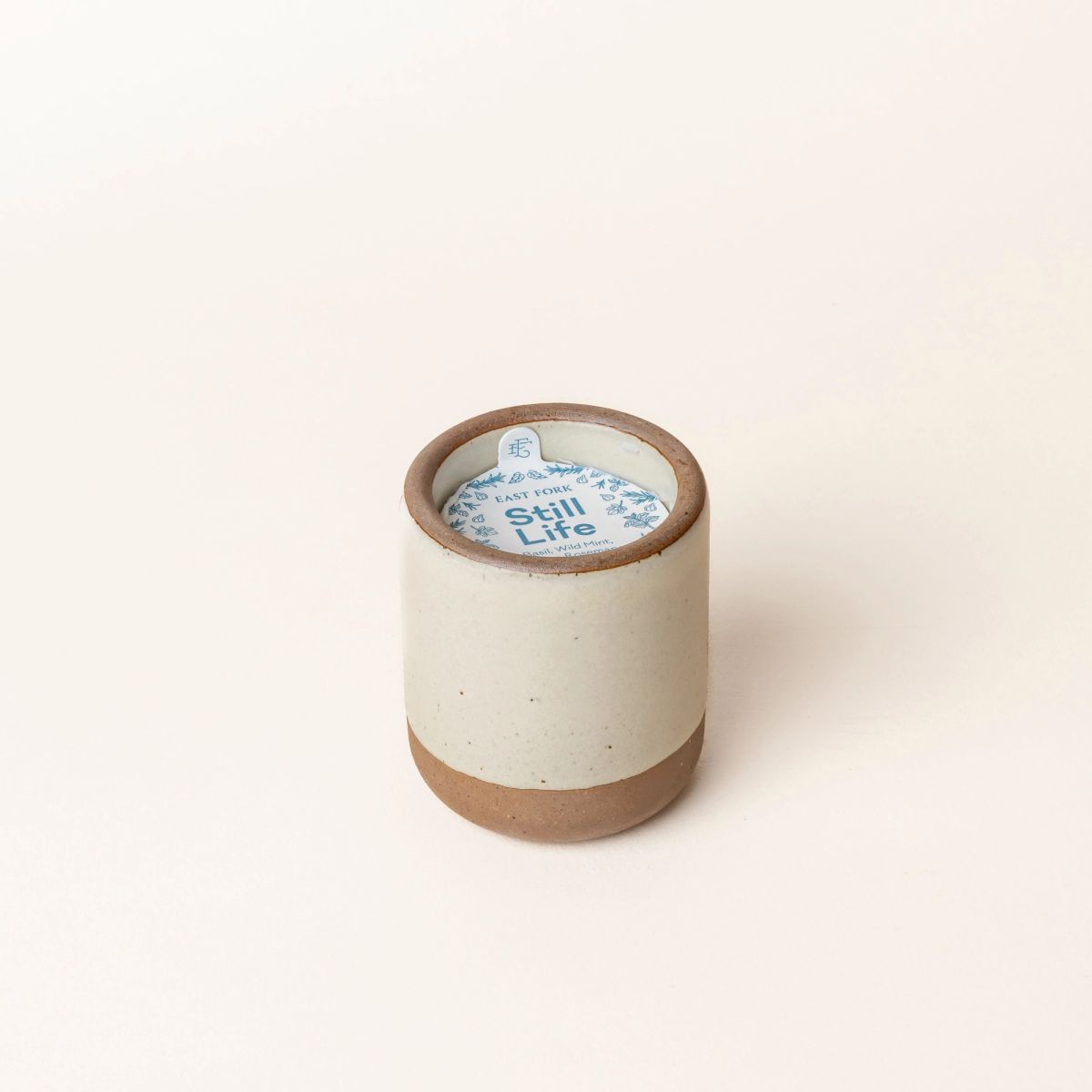 Small ceramic vessel in  warm, tan-toned, off-white color with candle inside. On top is a packaging label sitting on top that reads "Still Life"