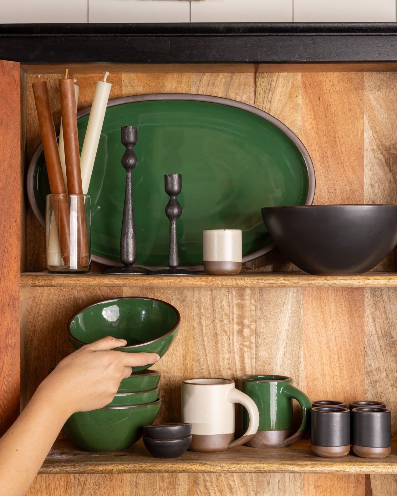 A hand places a glossy emerald green bowl on a stack on a shelf in a cabinet with more ceramic dinnerware, candlesticks, and iron candleholders