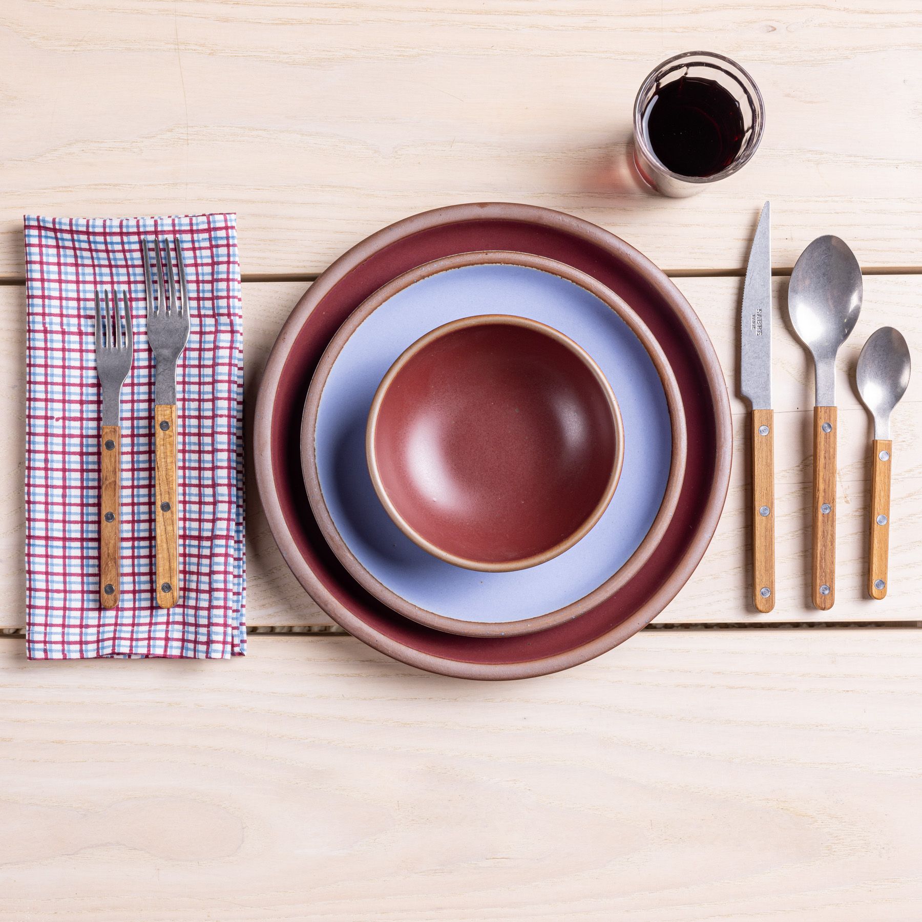 A table setting featuring teak-handled flatware, a juice glass, ceramic plates and bowl in periwinkle and plum, and a napkin.