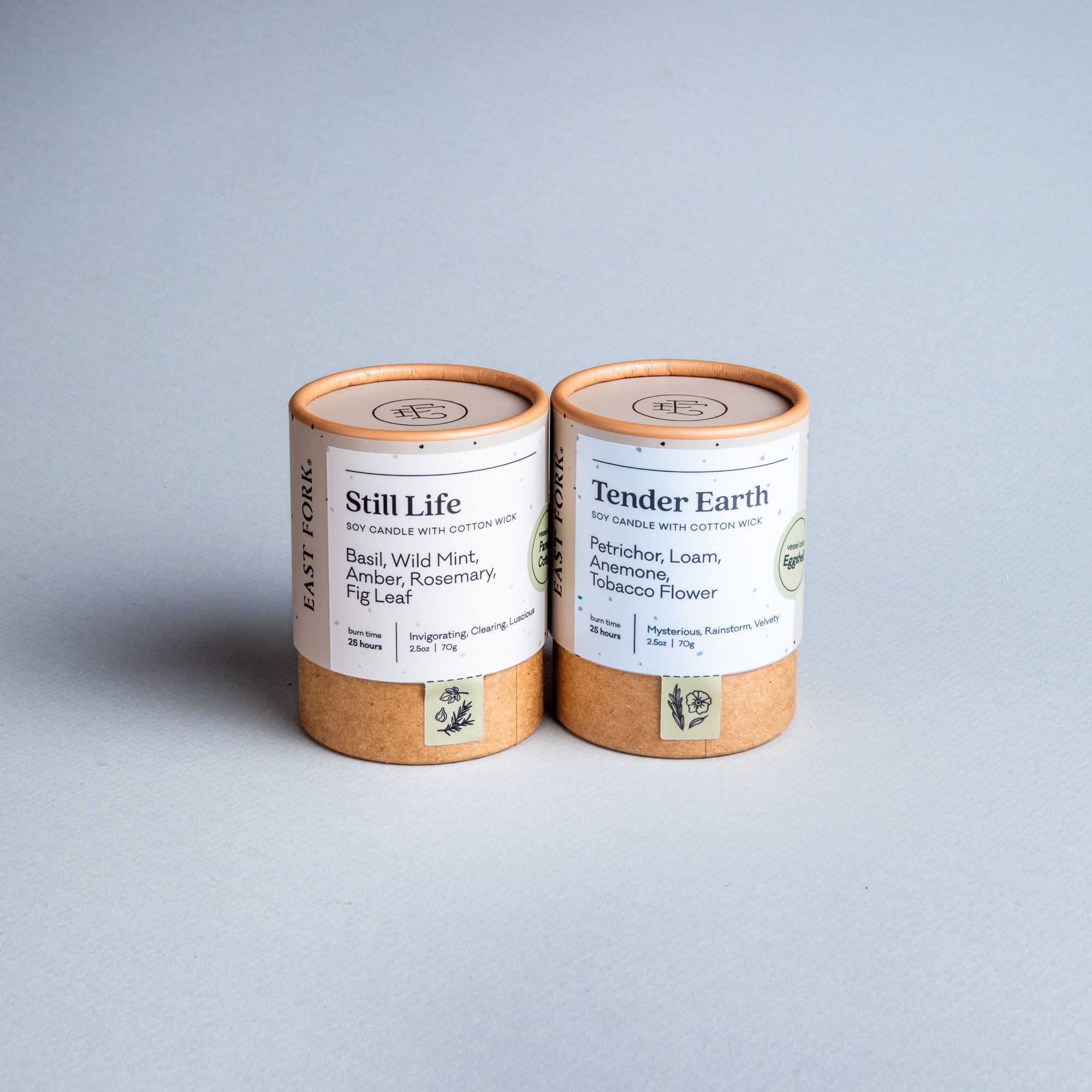 Two small cardboard packaging tubes with a candle inside each with branding on the outside that says 'Still Life' and 'Tender Earth'