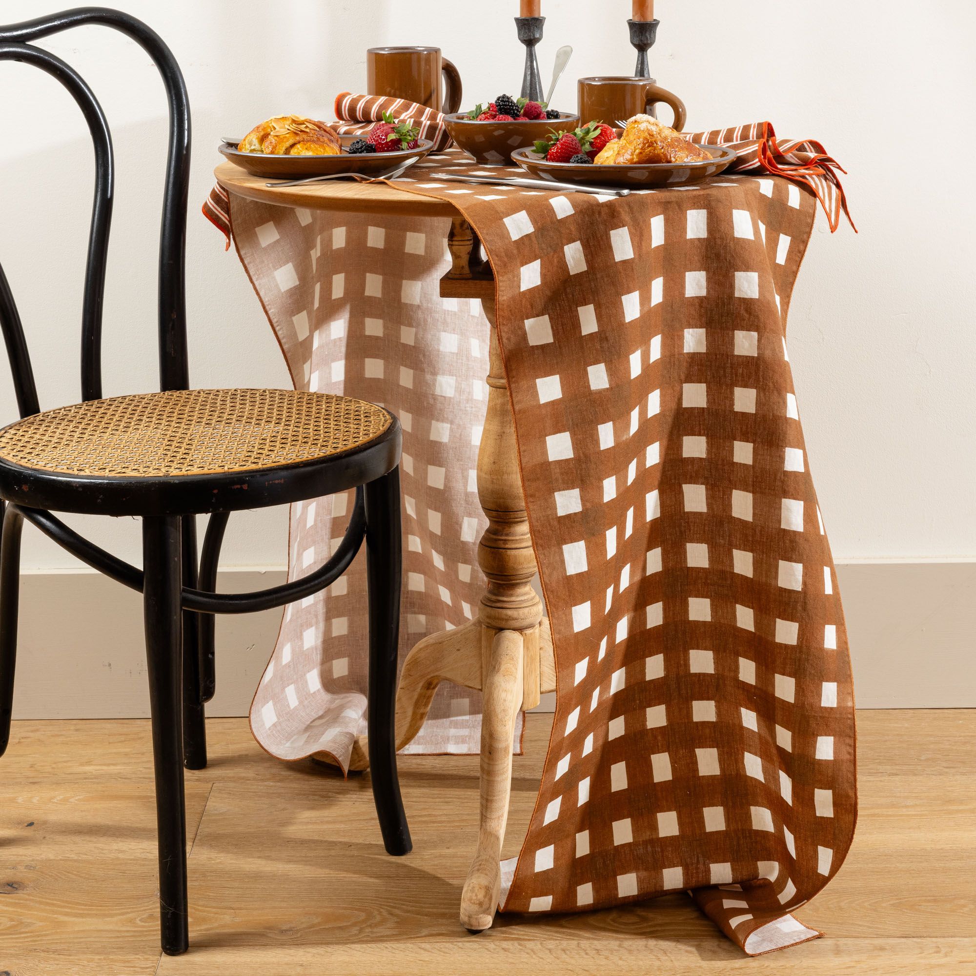 Brown crosshatch table runner draped over a small table set with breakfast pastries, fruit, and mugs, beside a black chair.