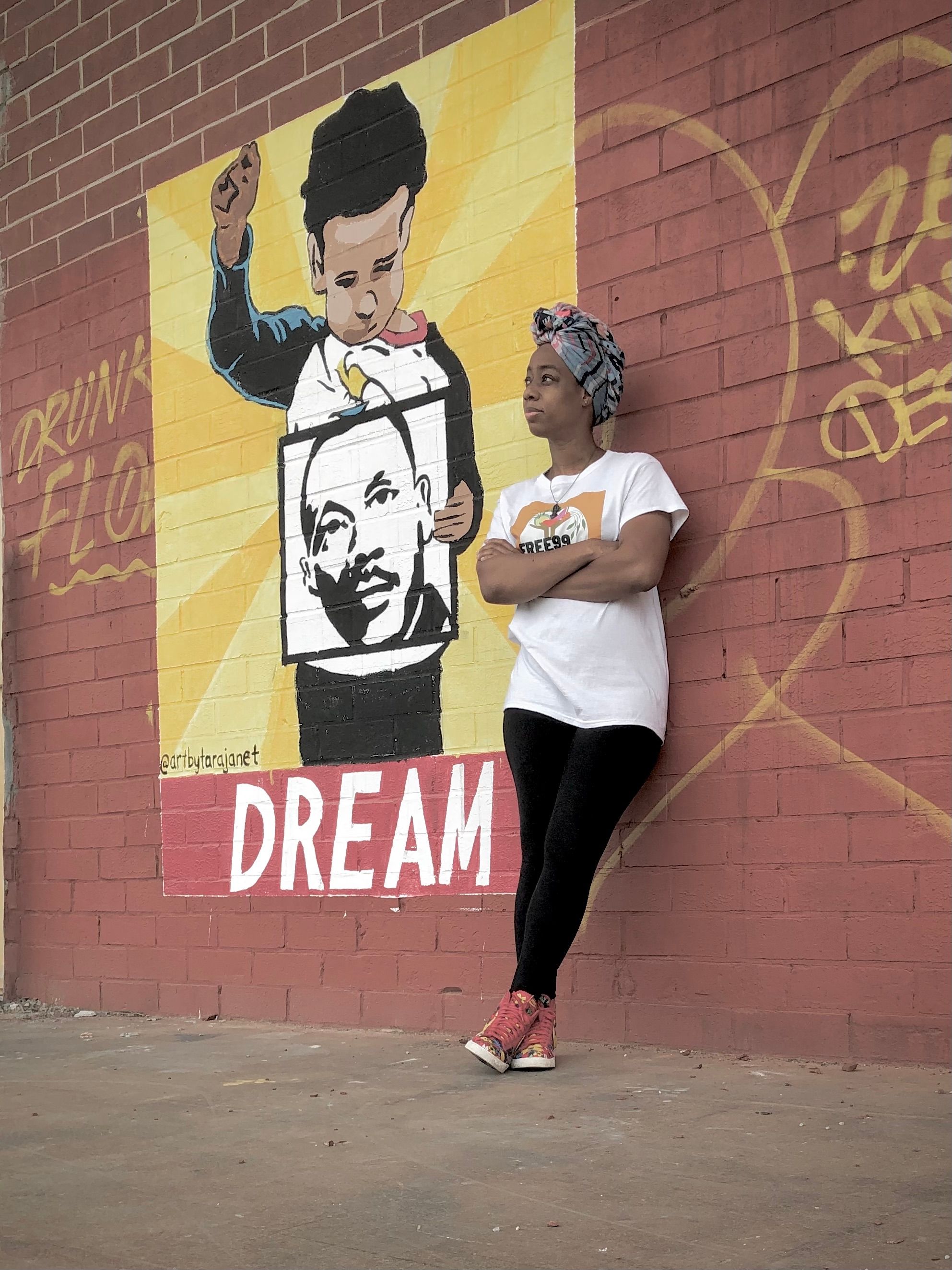 A person wearing black pants and a white graphic t-shirt leans against a brick wall with a mural of a child holding a Dr. Martin Luther King Jr. picture with the word "Dream" below.