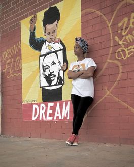 A person wearing black pants and a white graphic t-shirt leans against a brick wall with a mural of a child holding a Dr. Martin Luther King Jr. picture with the word "Dream" below.
