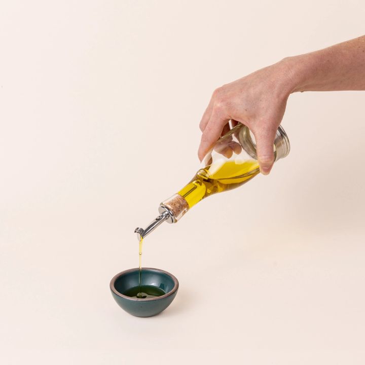 A hand holds a glass cruet that is pouring oil into a small dark teal tiny bowl. The cruet has a silver pewter base, glass body, and silver pourer and cork stopper. The cruet is wider at the bottom and narrows halfway to the the top.