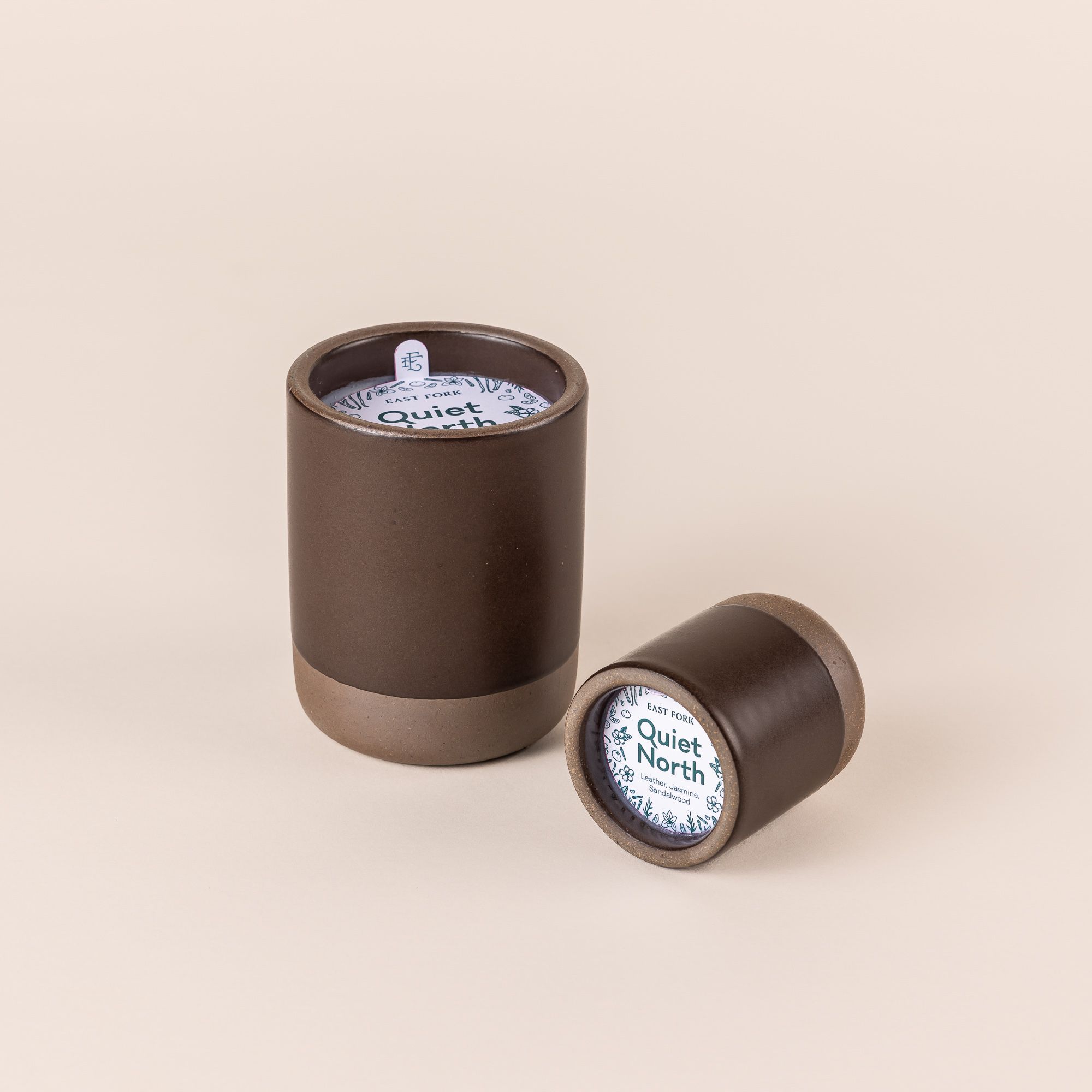 Large and small ceramic vessel in a dark cool brown color with candle inside. On top of each is a packaging label sitting on top that reads "Quiet North".