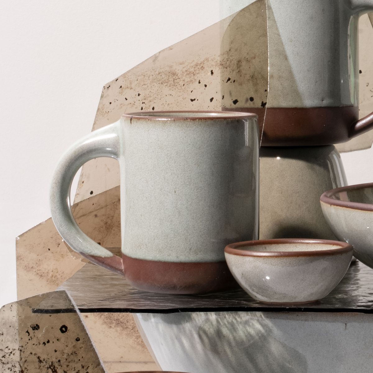Close-up of a reflective icy white ceramic mug and small bowl with unglazed rims, arranged with reflective textured glass panels in a minimalist setting.