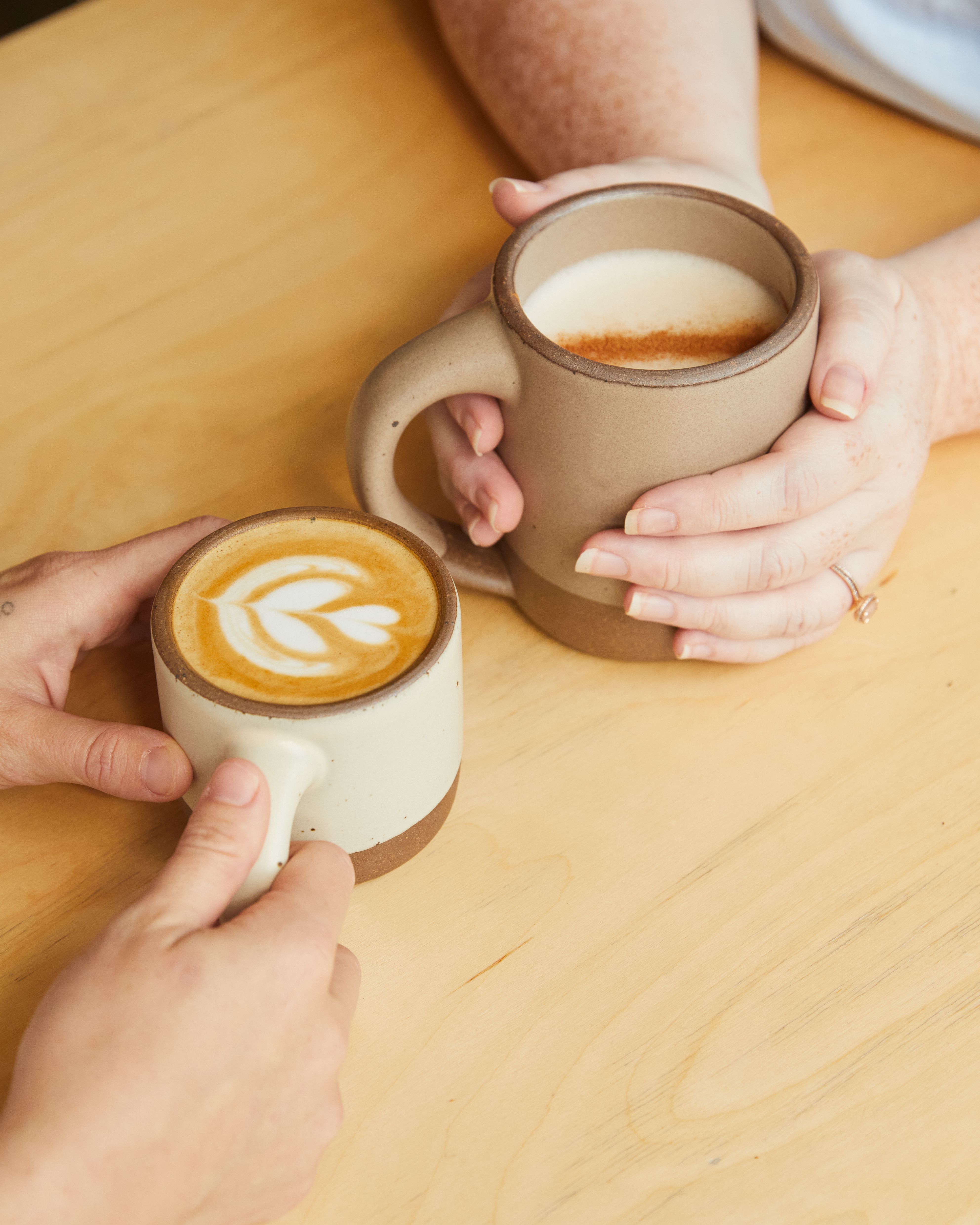 Two sets of hands are holding ceramic mugs filled with coffee at a wooden table