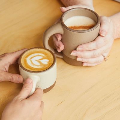 Two sets of hands are holding ceramic mugs filled with coffee at a wooden table