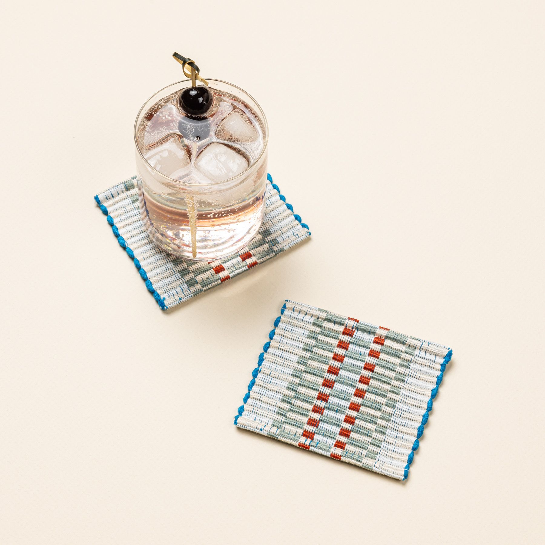 A cocktail sits on 1 of 2 square coasters that are hand woven in a striped rectangular grid design in sage, orange, and blue colors