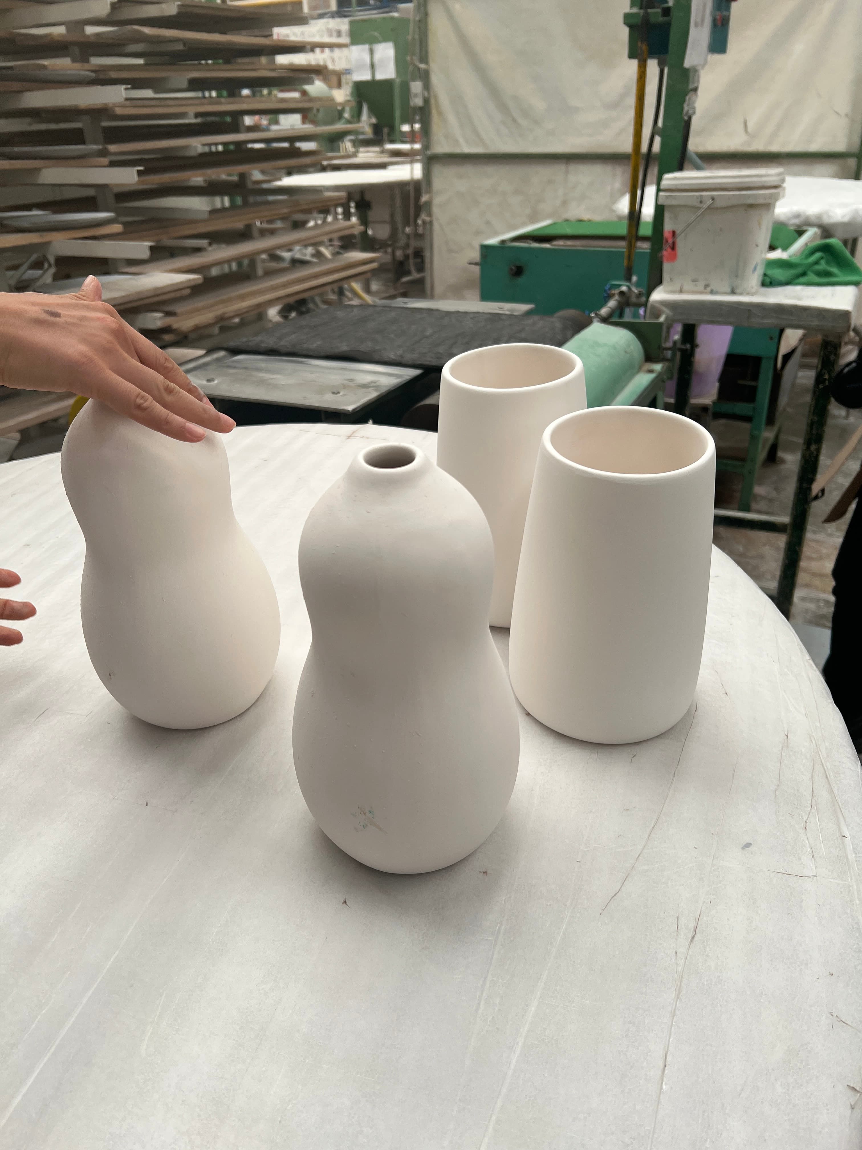 A table in a pottery studio with four unglazed ceramic vases—two gourd-shaped and two cylindrical—being inspected by a person’s hand. Shelves, tools, and other pottery equipment are visible in the background.