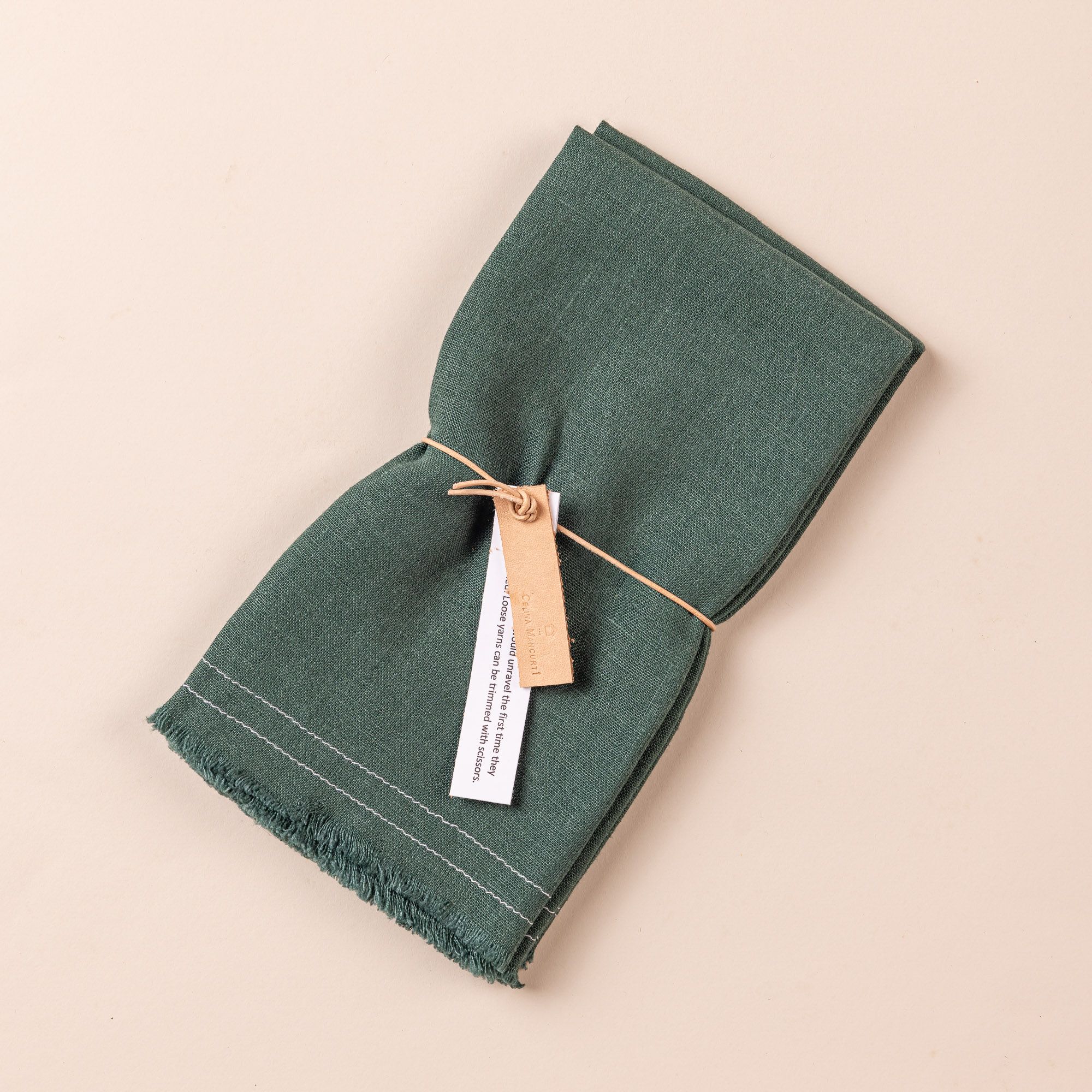 A folded deep teal pair of linen napkins with a string and small tag wrapped around the center. The bottom end of the napkins has a fringe edge.