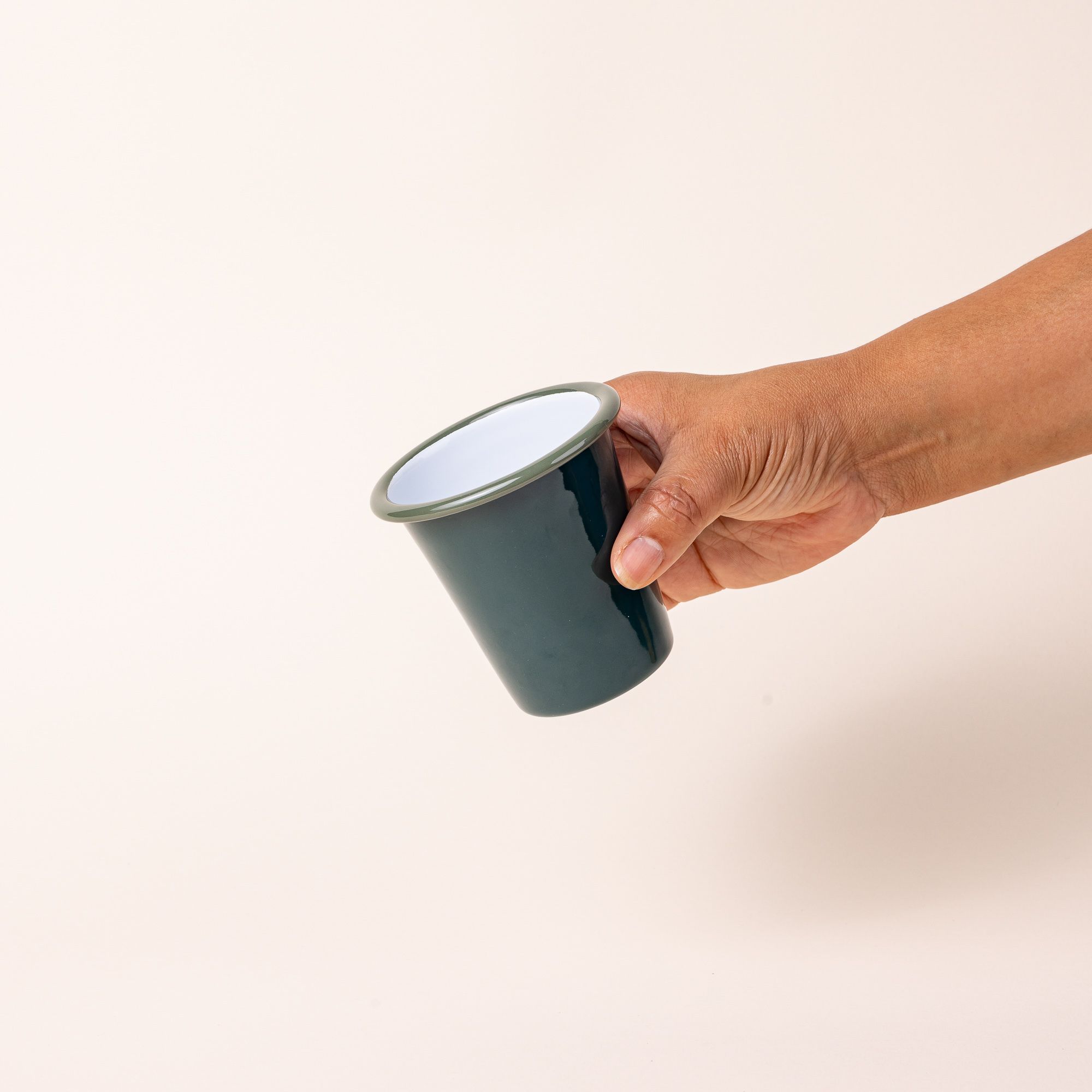 A hand holding a short enamel cup with dark green teal exterior and white interior