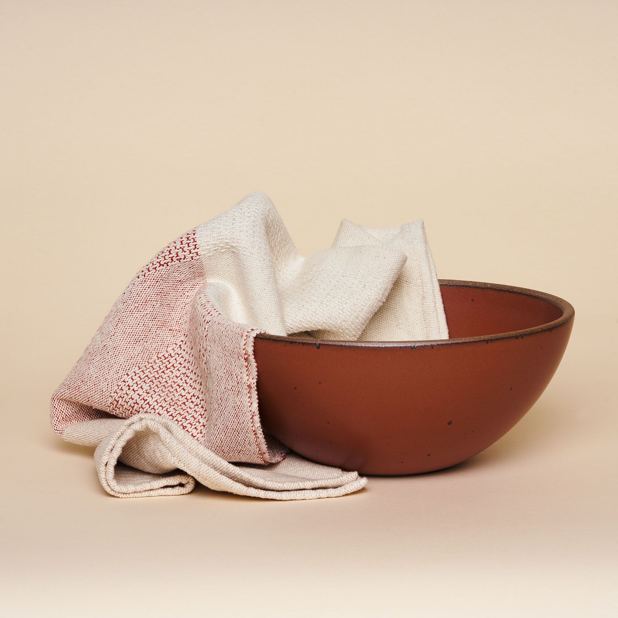 A half-red, half-natural woven tea towel placed whimsically in a matching earthy red stoneware pottery bowl.