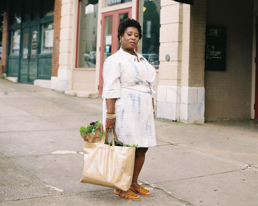 A woman stands on a city block wearing a dress and holding a tote bag of a farmer's market haul.