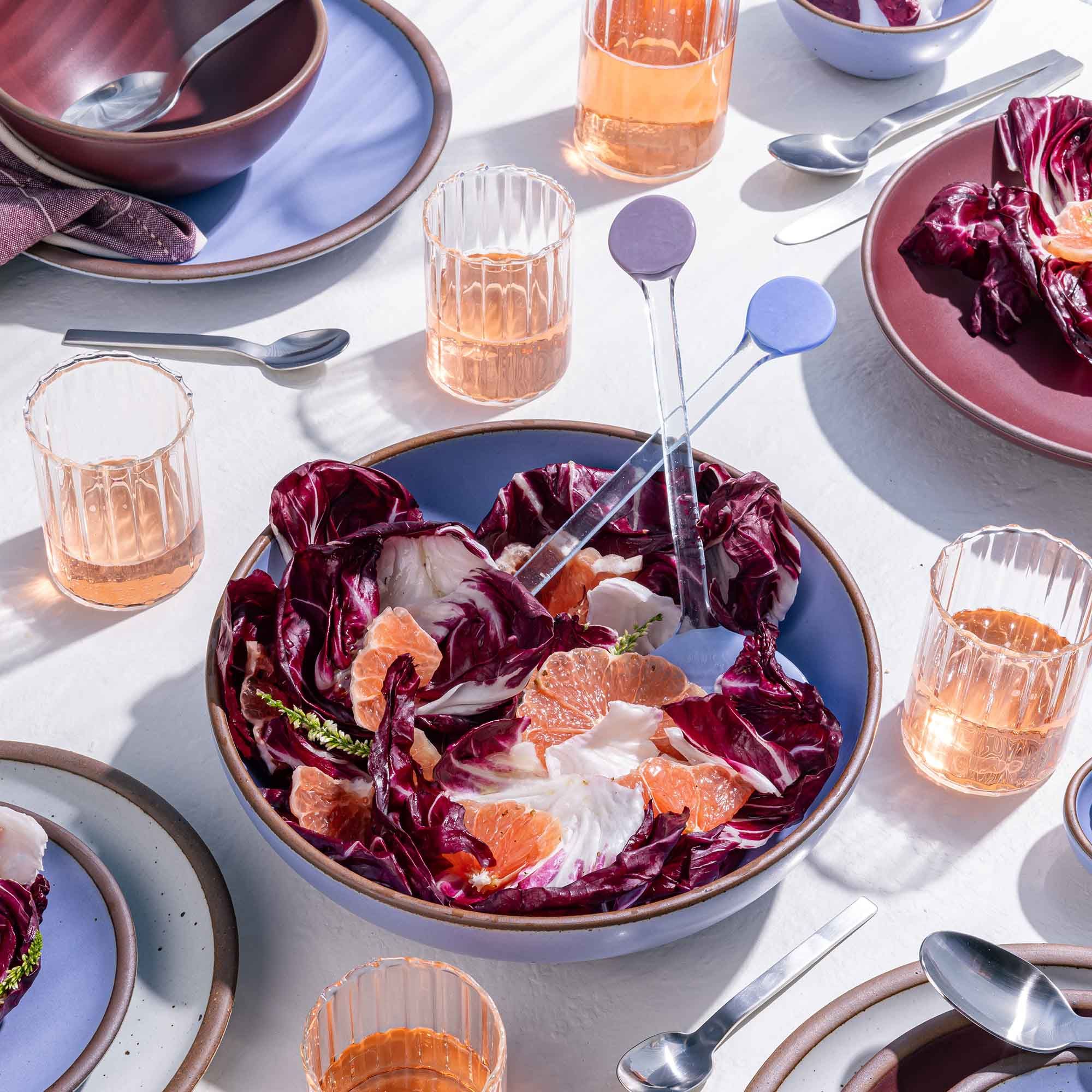 A table with a serving bowl filled with salad and glass serving spoons, with glasses and plates.