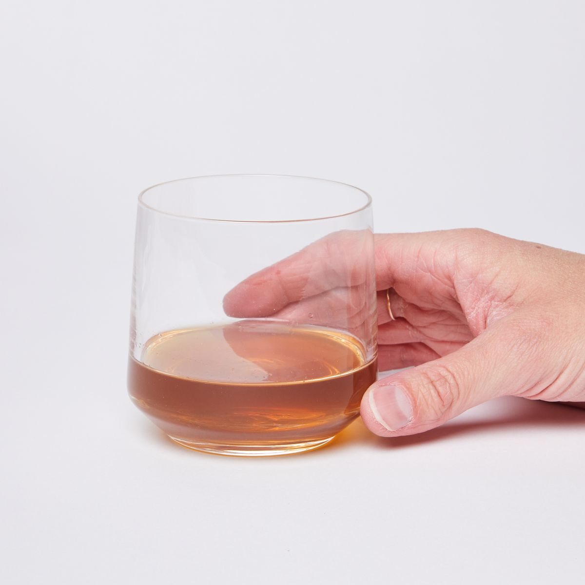 Hand holding a clear glass tumbler with a small pour of brown liquid