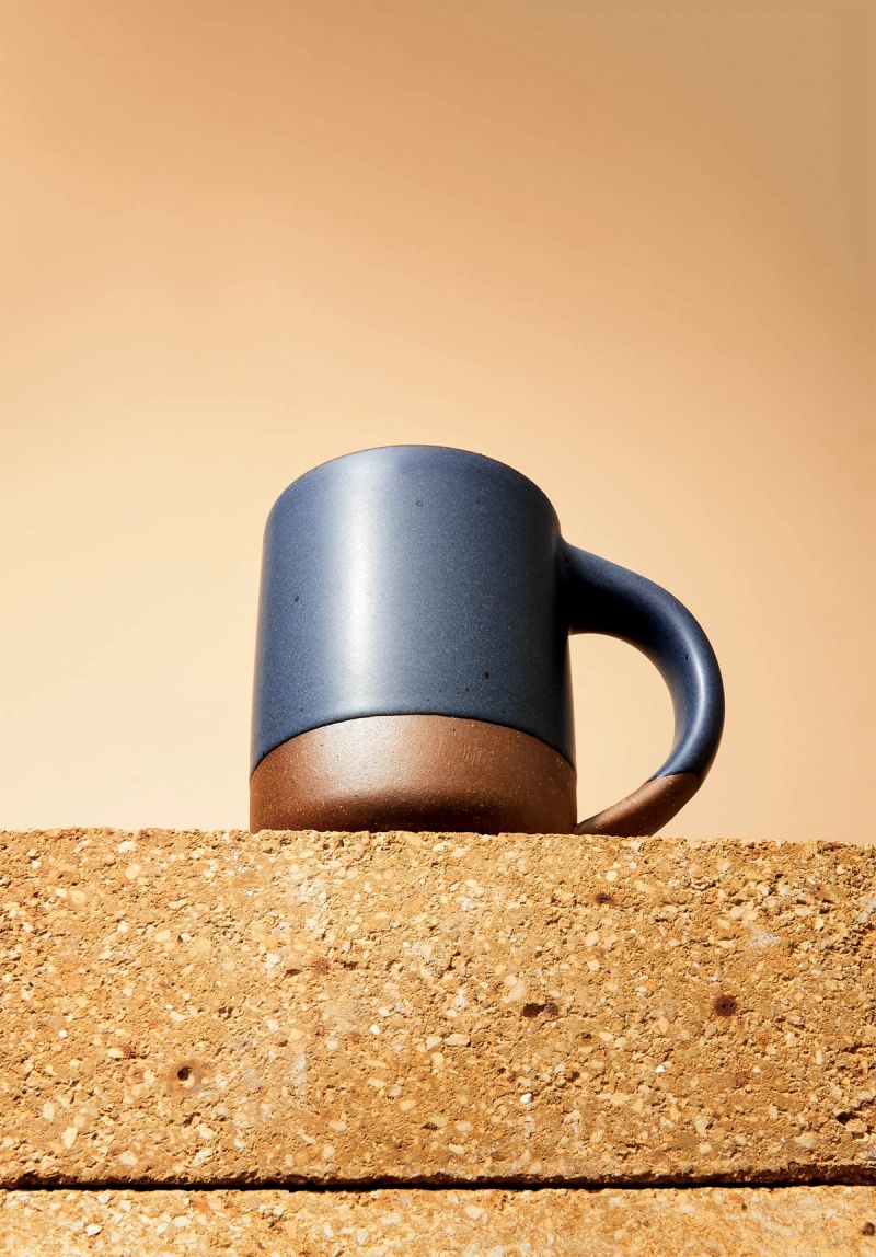 A ceramic mug sits on a textured riser with the camera angled to look up at it from the bottom. The mug is ceramic and features a muted navy color and a raw glaze bottom.