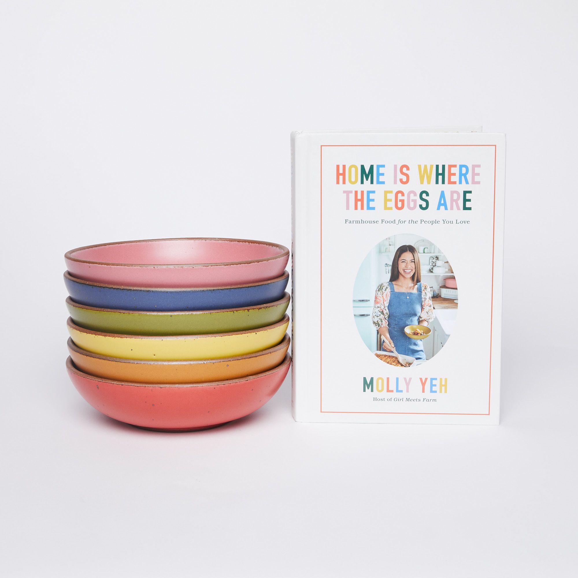 Cookbook next to stack of everyday bowls, from bottom to top - red, orange, yellow, green, blue, pink