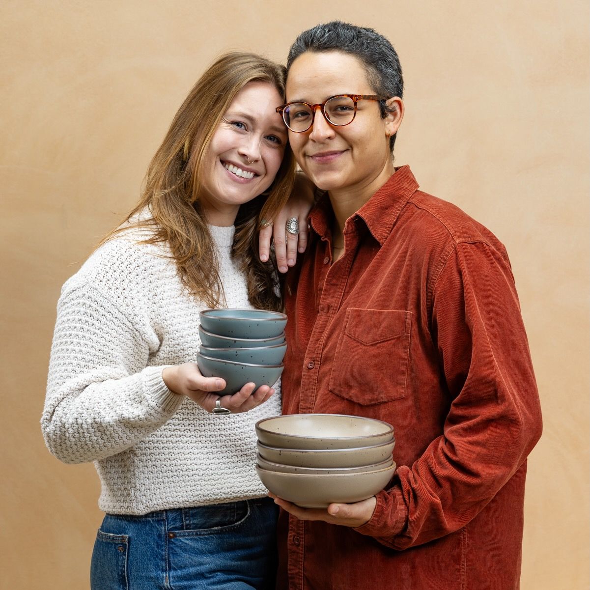 A couple is smiling and close together holding stacks of ceramic bowls in a muted tan and blue-grey colors.
