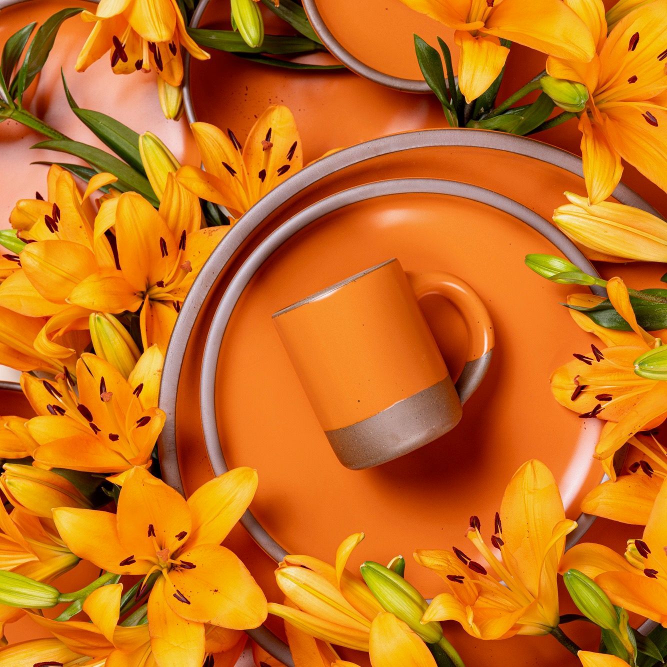 A close up of ceramic plates and a mug in a bold orange surrounded by blooming daylily flowers.
