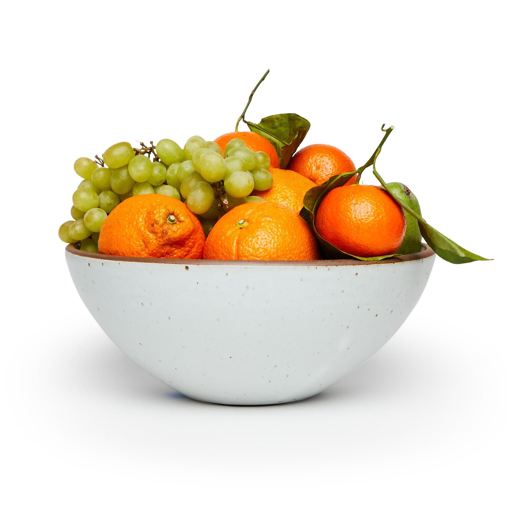 A large ceramic mixing bowl in a cool white color featuring iron speckles and an unglazed rim filled with a medley of fruit
