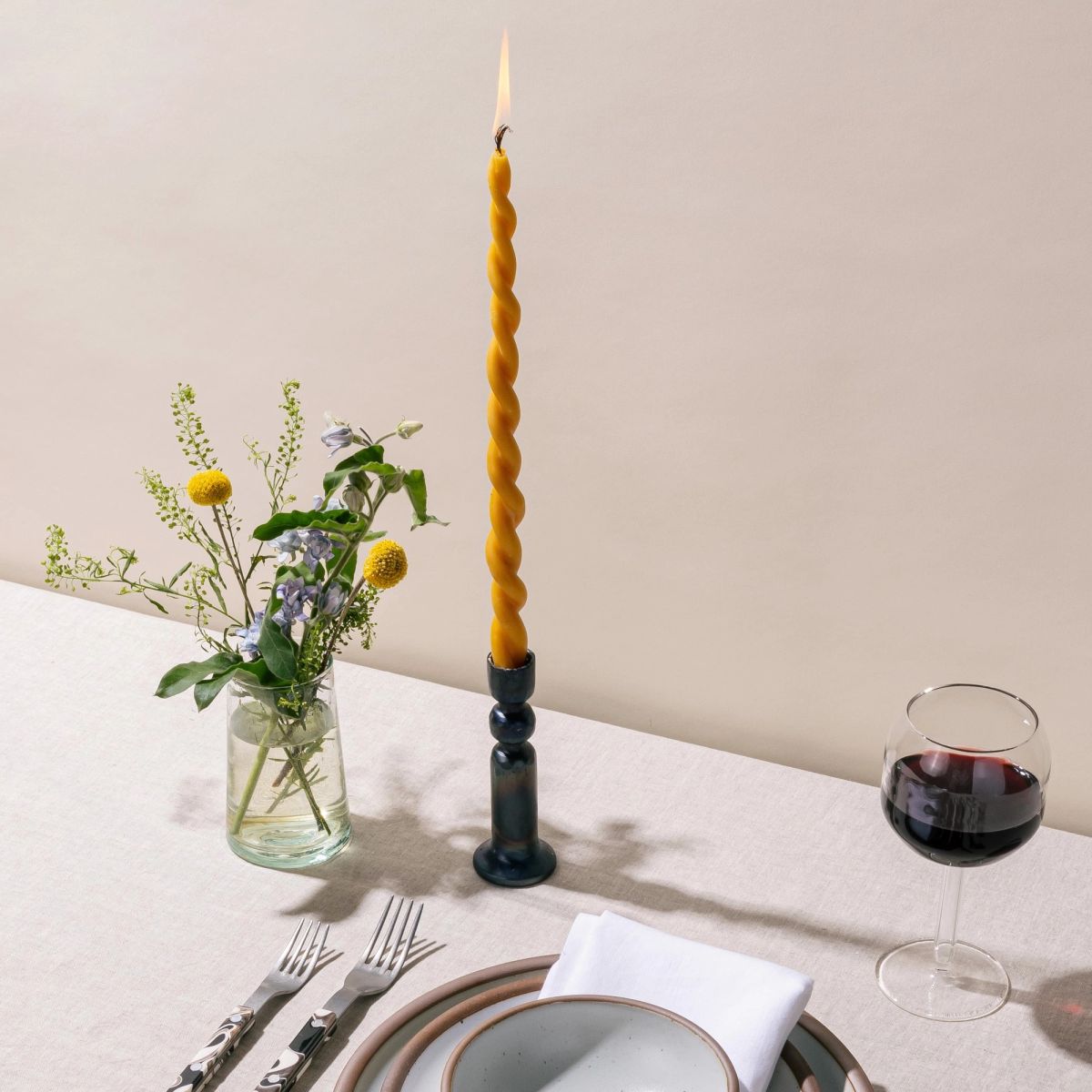 A iron candlestick holder with a lit twisted beeswax taper candle, a glass of flowers, a glass of red wine and a table setting all sit on a table against a cream background.