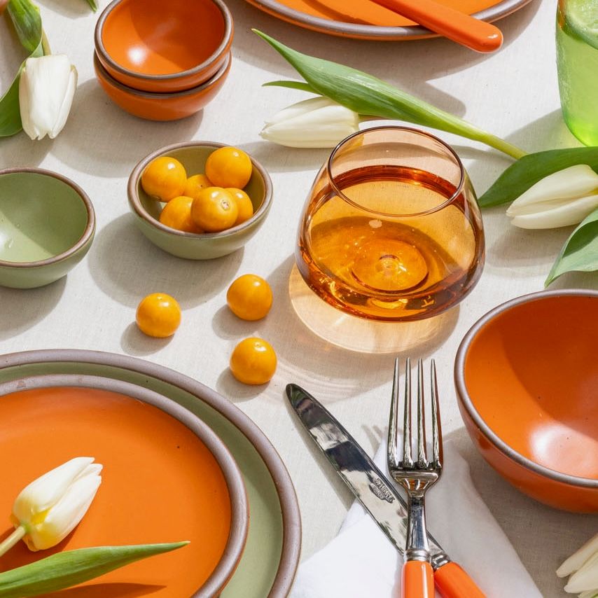 On a table is a colorful setting with an orange glass whiskey snifter, ceramic plates and bowls in orange and sage green colors, a fork and knife, and little orange tomatoes and tulips artfully arranged.