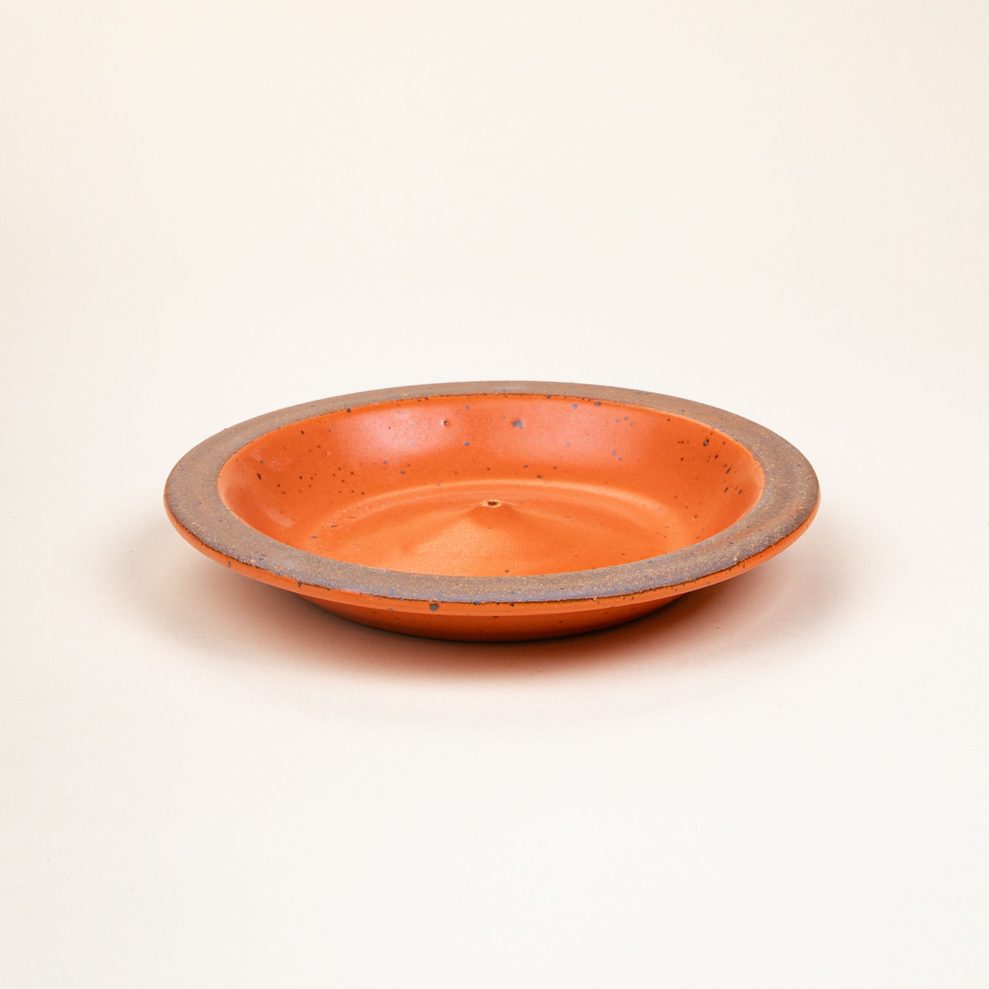 A ceramic plate-shaped incense stick holder with an unglazed rim and glazed in a bold orange color featuring lots of iron speckles.