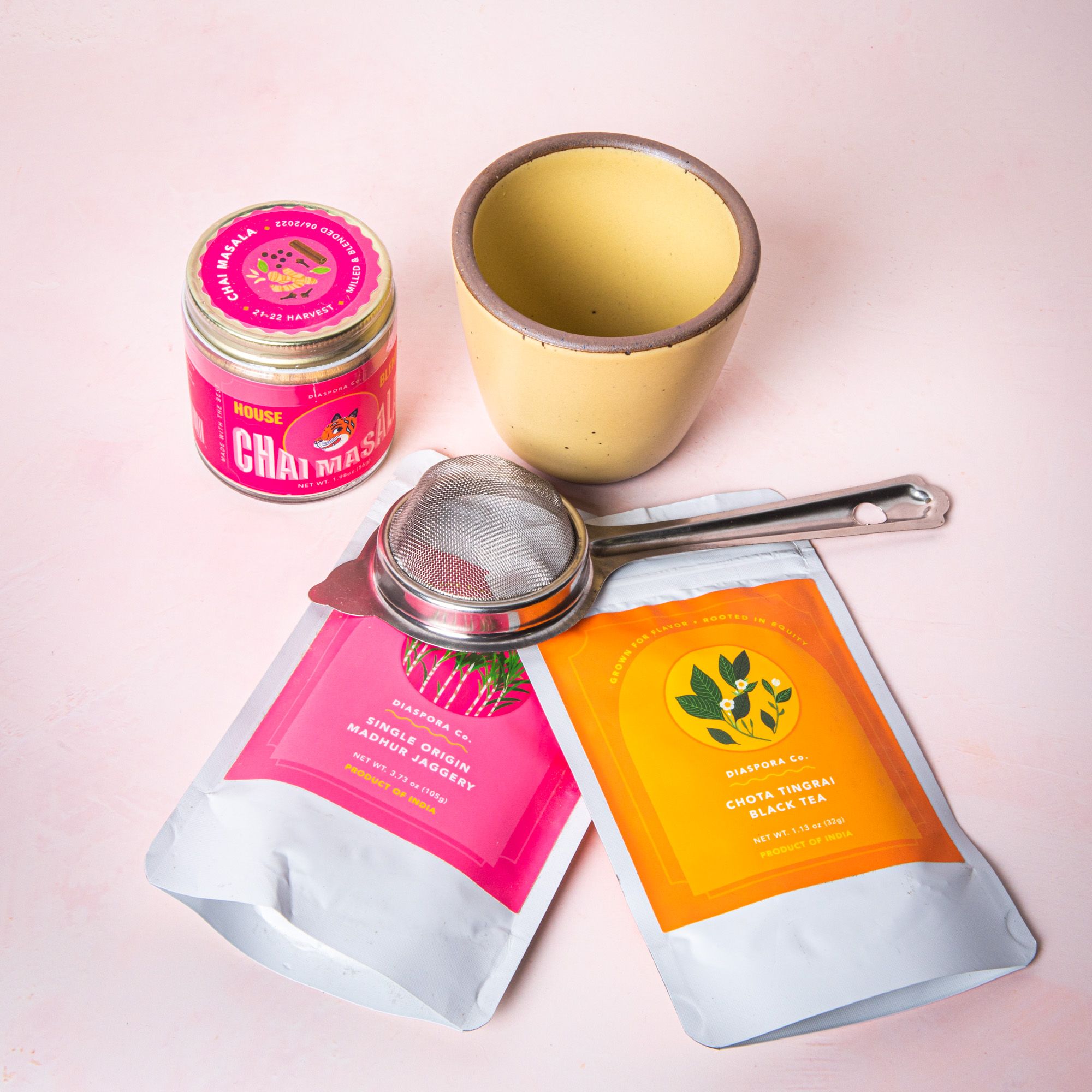 A chai bundle on a soft pink background featuring a soft butter yellow kulhad, a tea strainer, a pink jar of chai masala, and 2 packages of jaggery and black tea