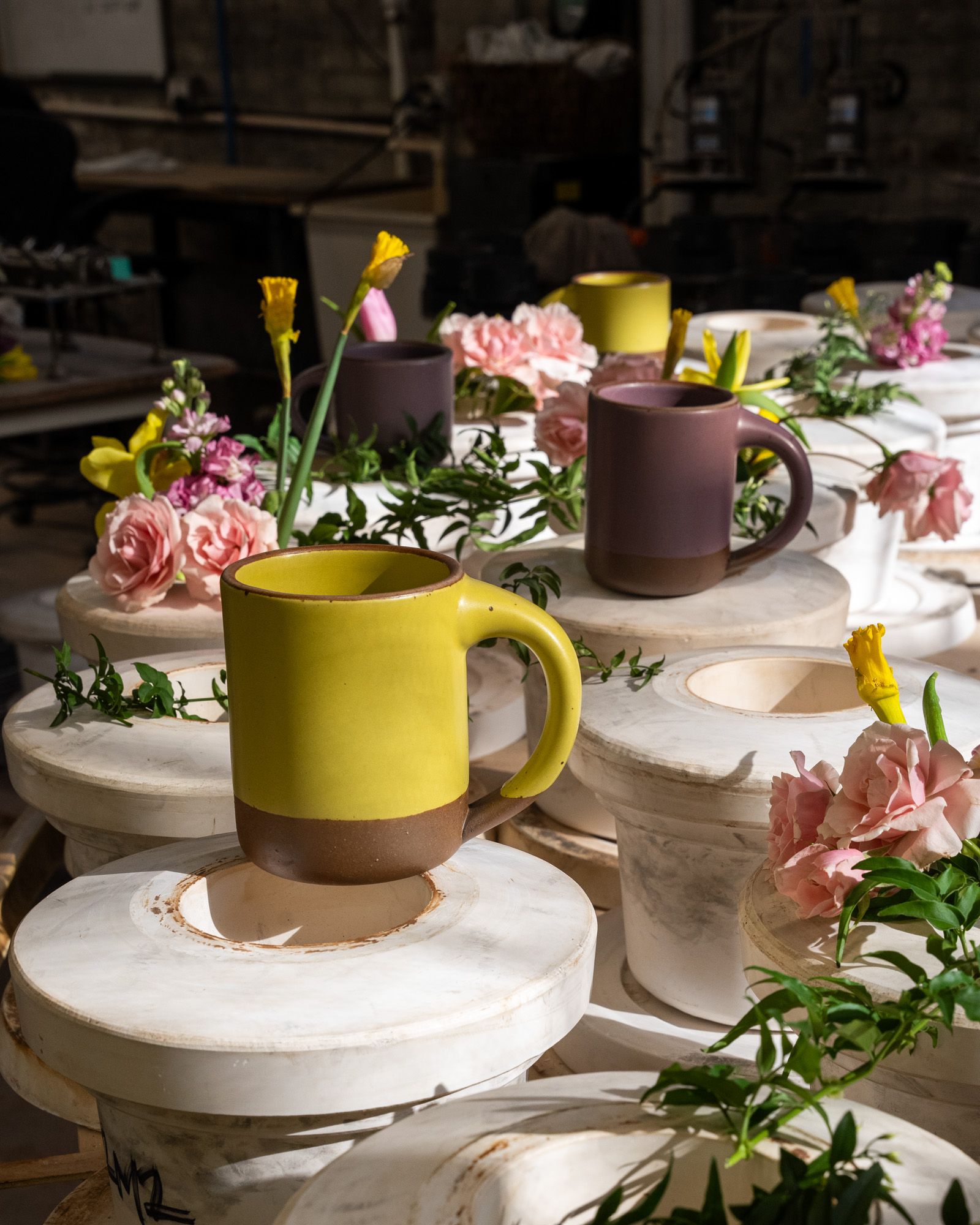 On a large table to form ceramic mugs, finished ceramic mugs in zesty yellow and soft mauve purple colors are artfully arranged with flowers.