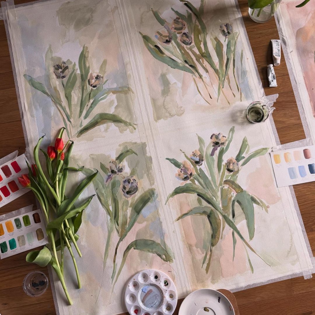 Watercolor painting of flowers along with flower stems, paint palettes and a paint cup of water.