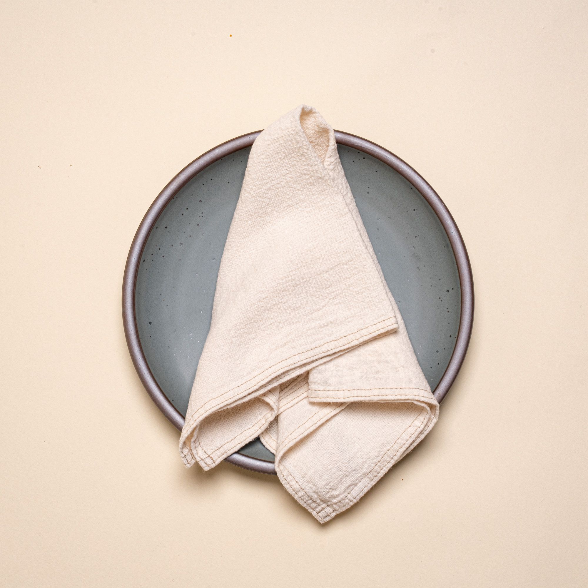 Folded cream linen napkin with sewn edge on a blue-grey ceramic dinner plate