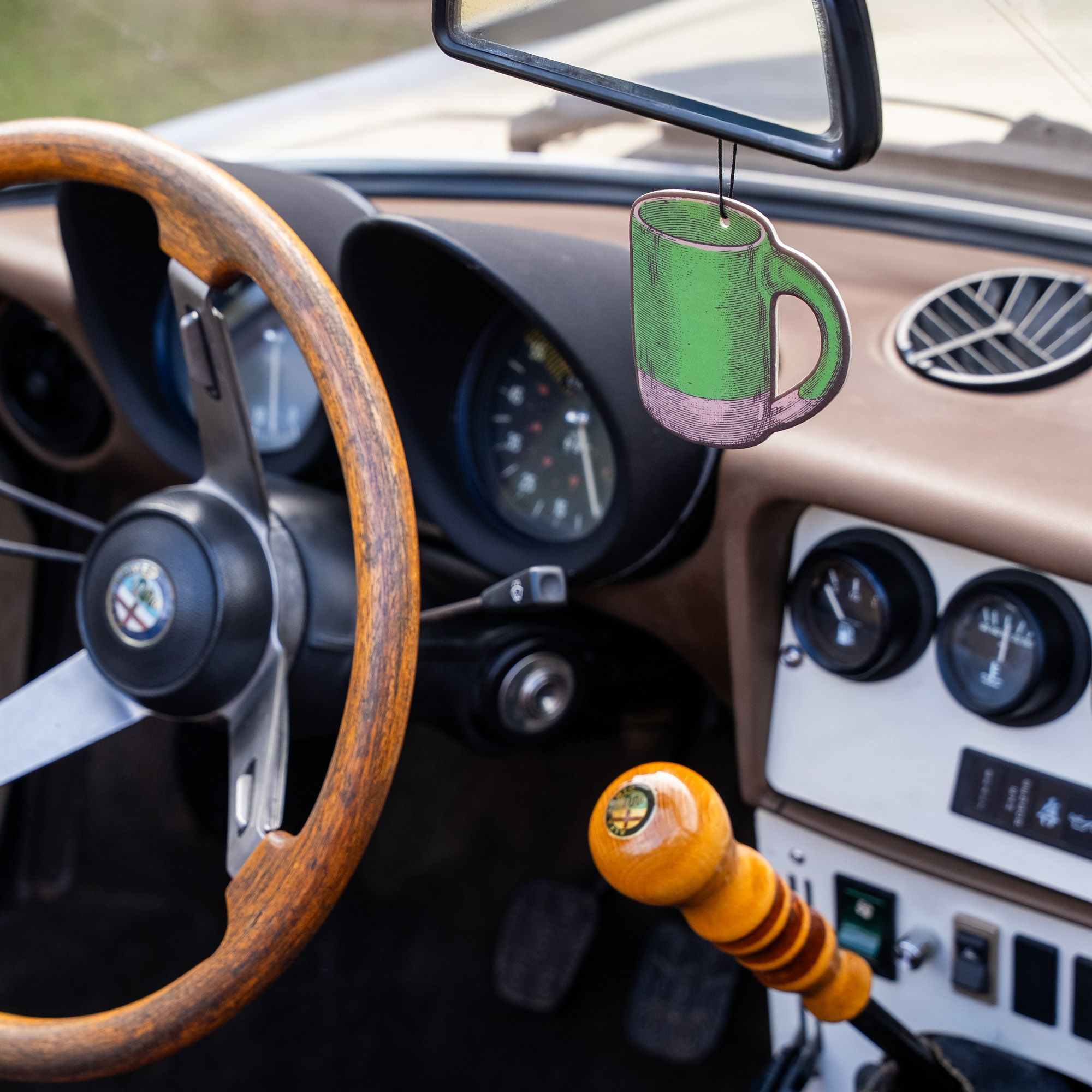 Green mug-shaped East Fork air freshener hanging from a rearview mirror inside a vintage car with a wood steering wheel and classic dashboard.