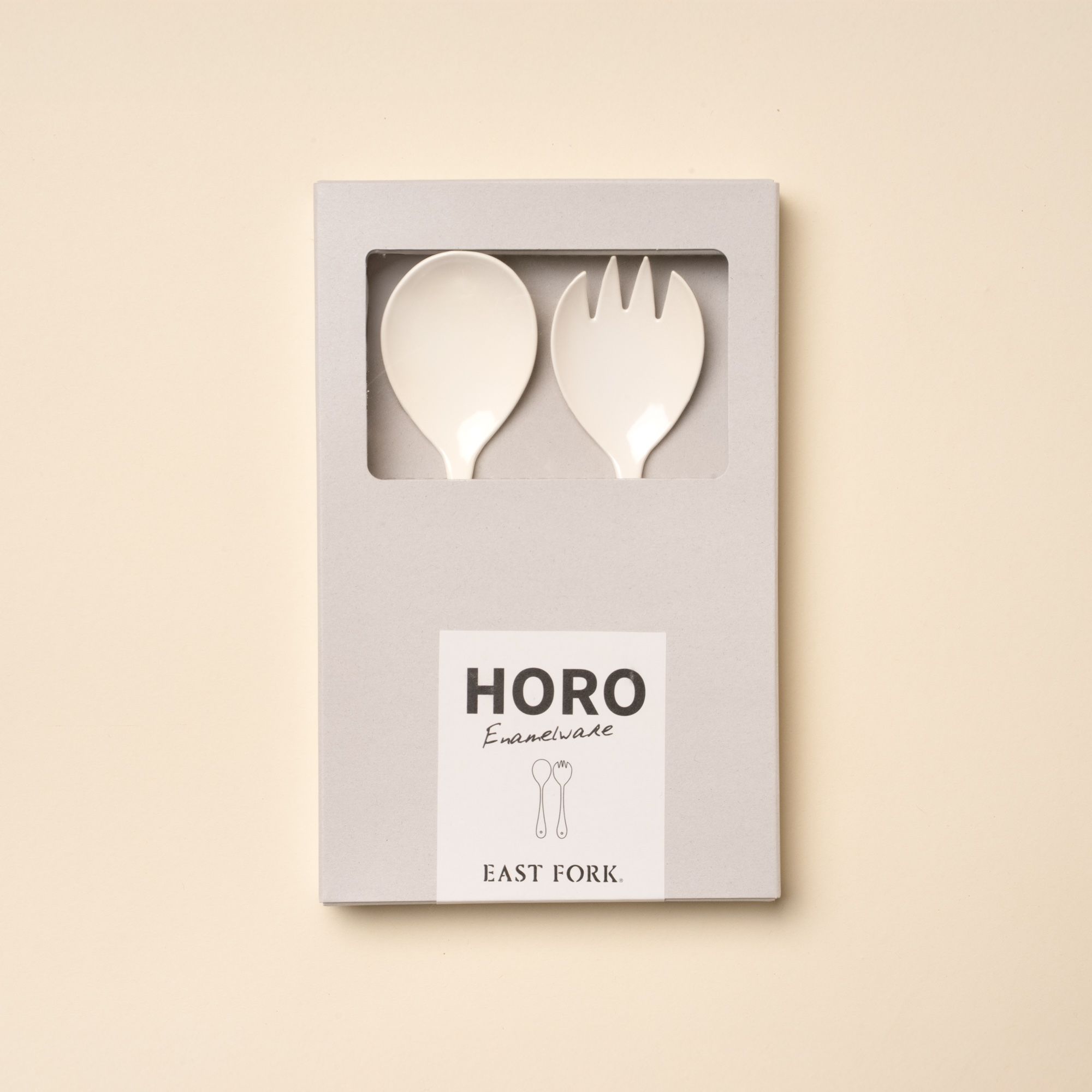 A serving fork and spoon set made of white enamel in a gray packaging box.